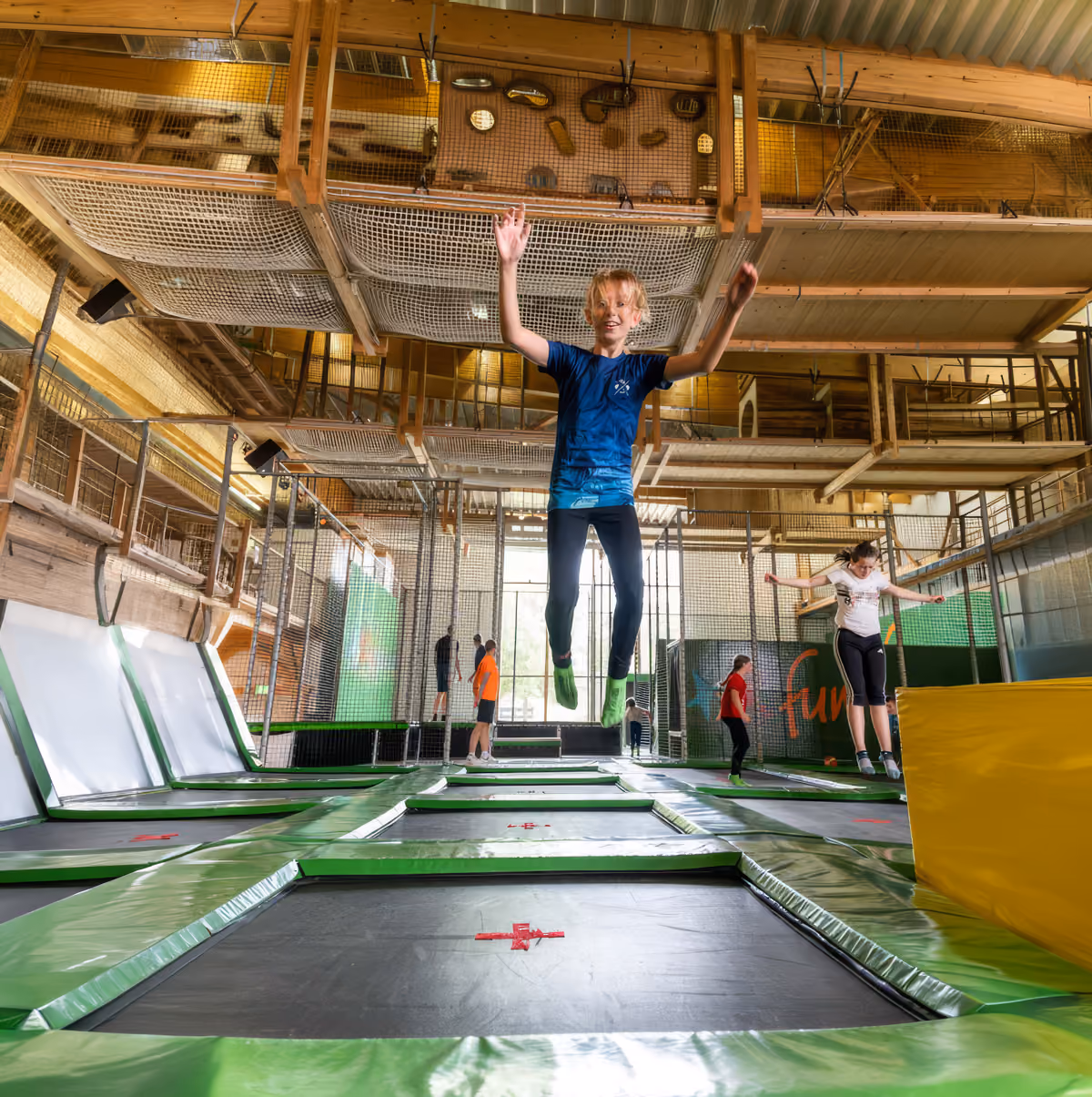 Kinder springen auf grünen Trampolinen in einer großen, hellen Indoor-Sprungarena.