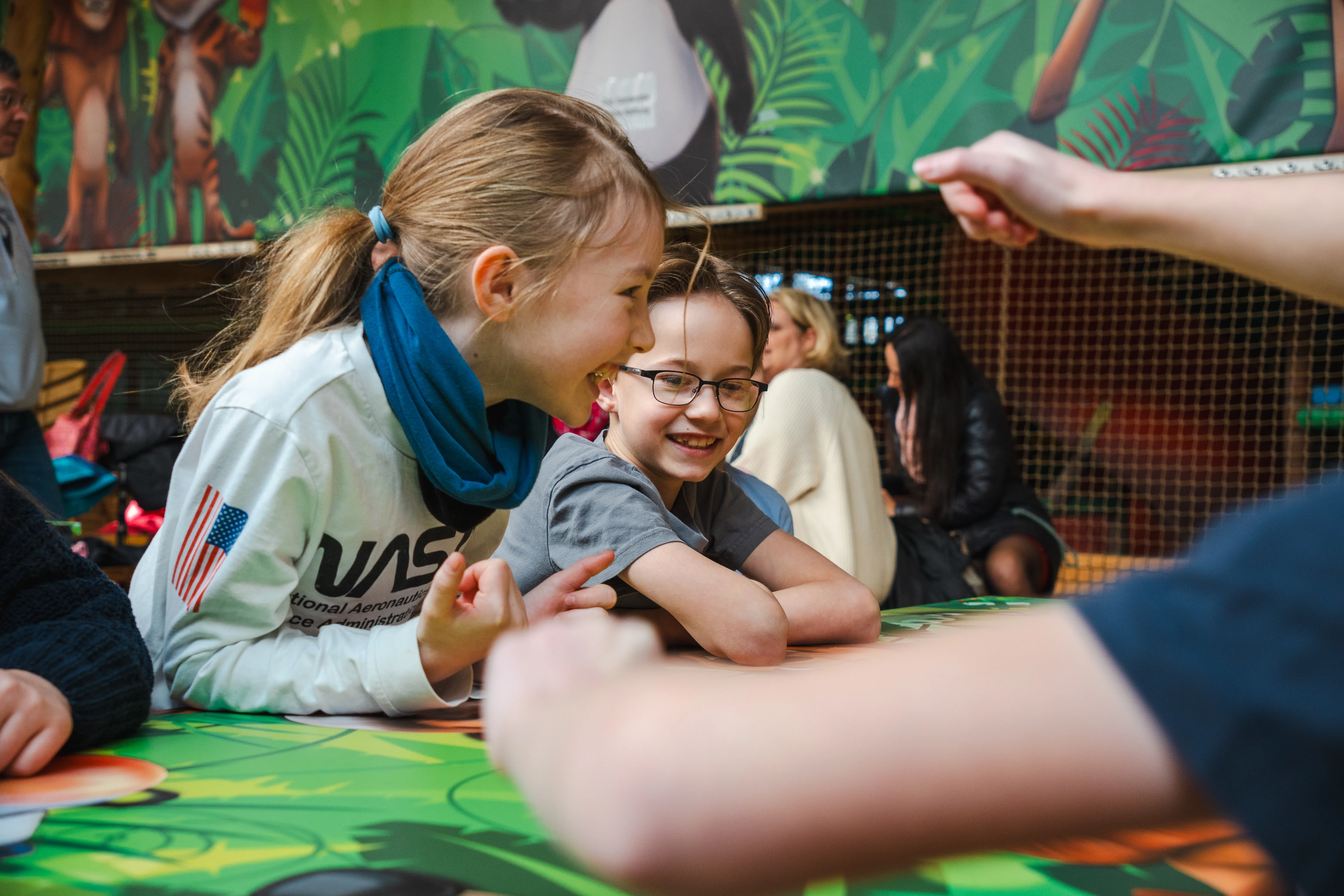 Zwei Kinder lachen und spielen an einem bunten Tisch in einem Raum mit Tiermotiven.