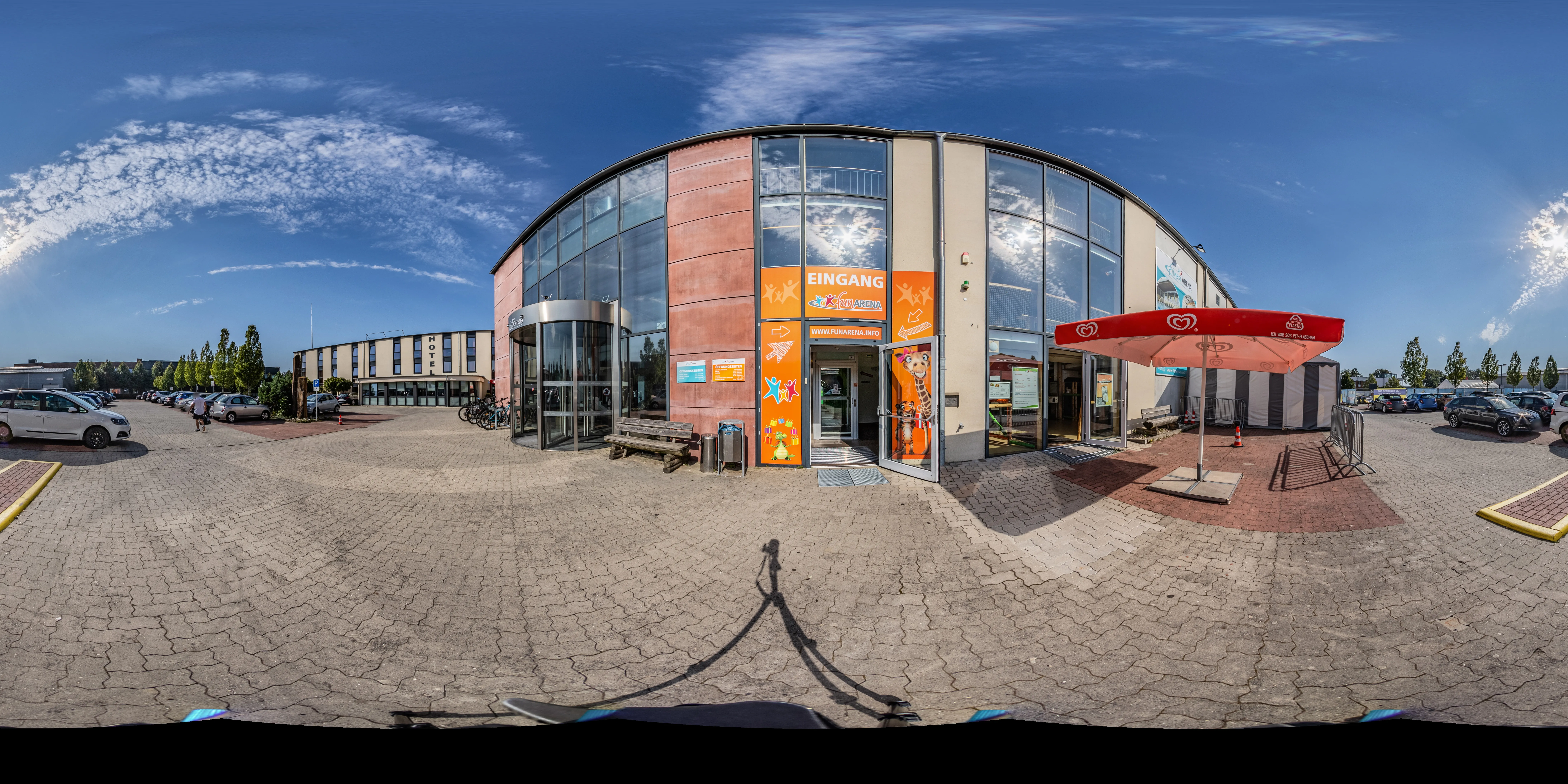 Eingang der FunArena Freizeit- und Sporthalle mit Parkplatz, rotem Sonnenschirm und blauem Himmel.