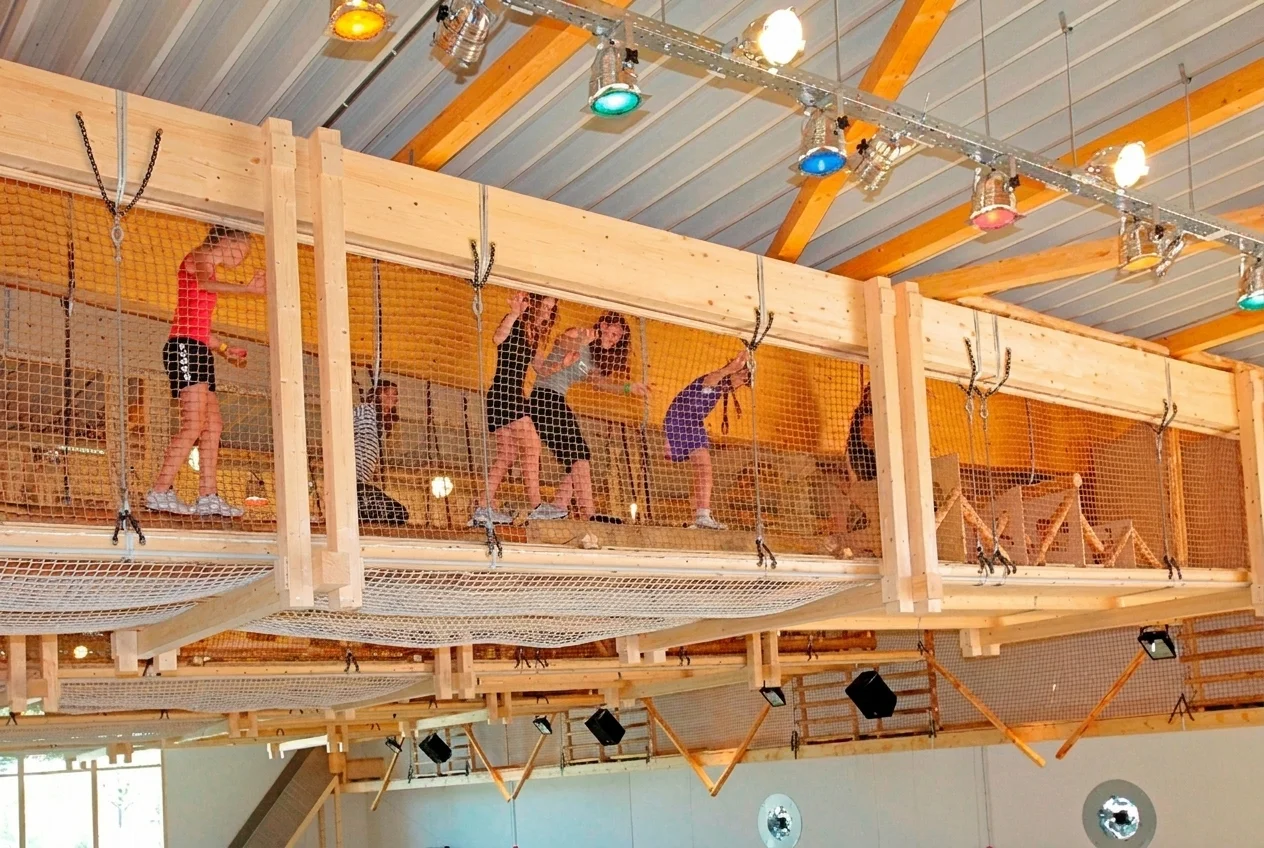 Kinder klettern auf einem erhöhten Holzgang mit Netzen in einer Innenhalle.