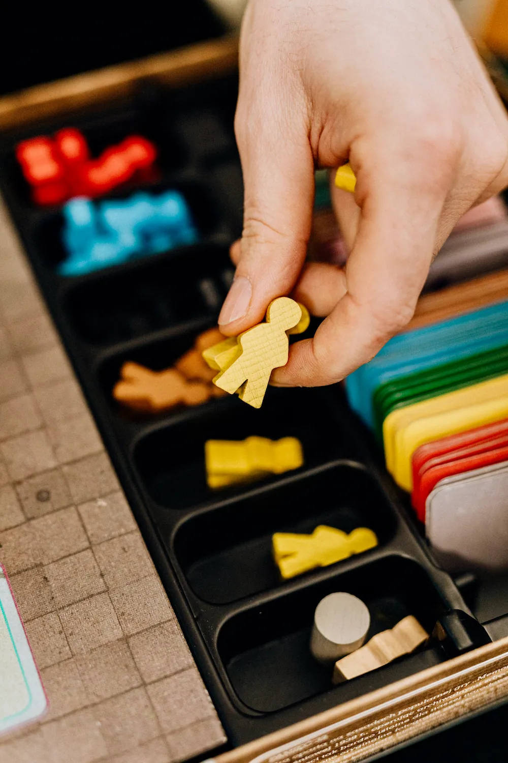 A hand picking a wooden meeple from a strategy board game's compartmented piece tray.