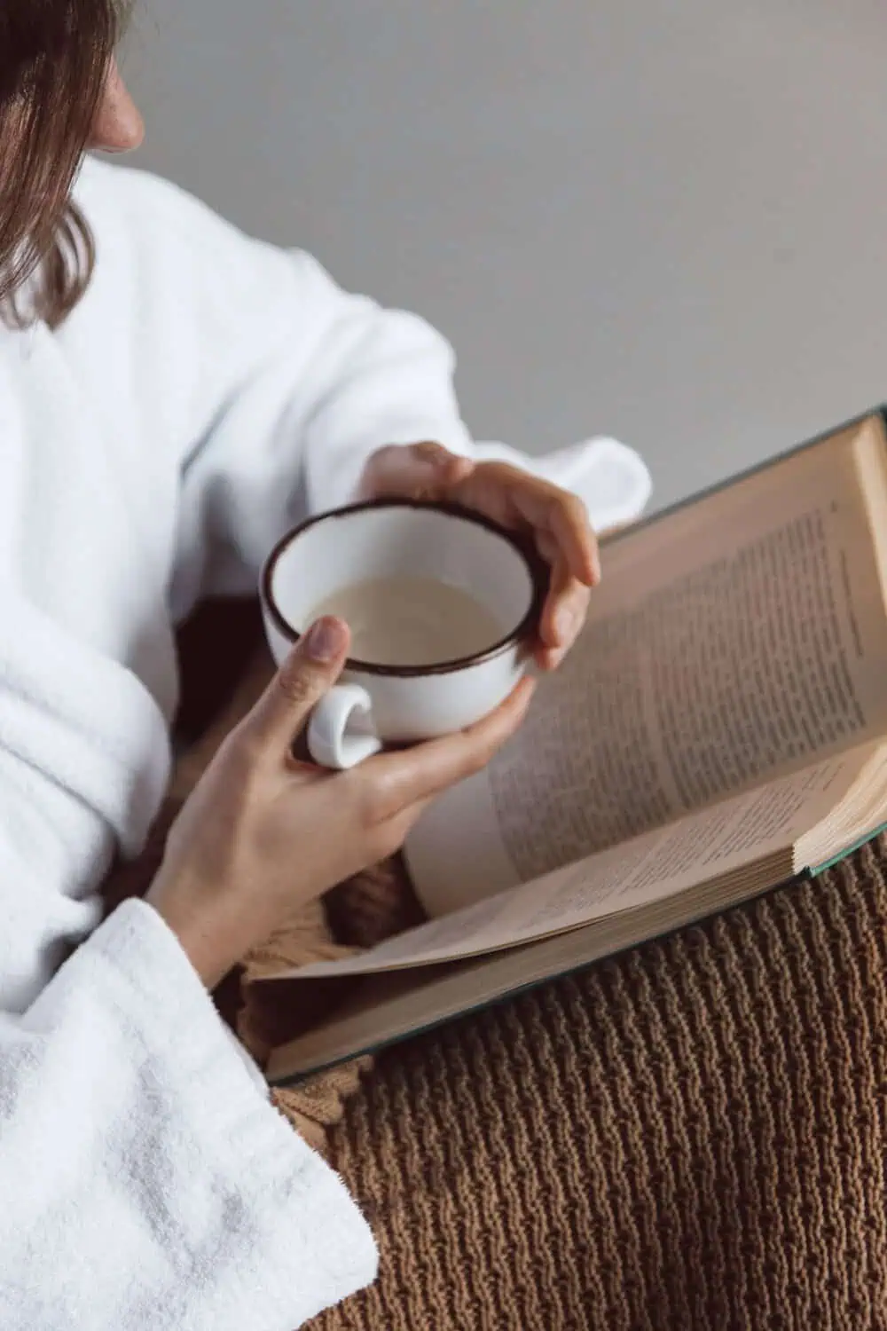 A person in a robe holding a mug while reading an open book, wrapped in a knit blanket.