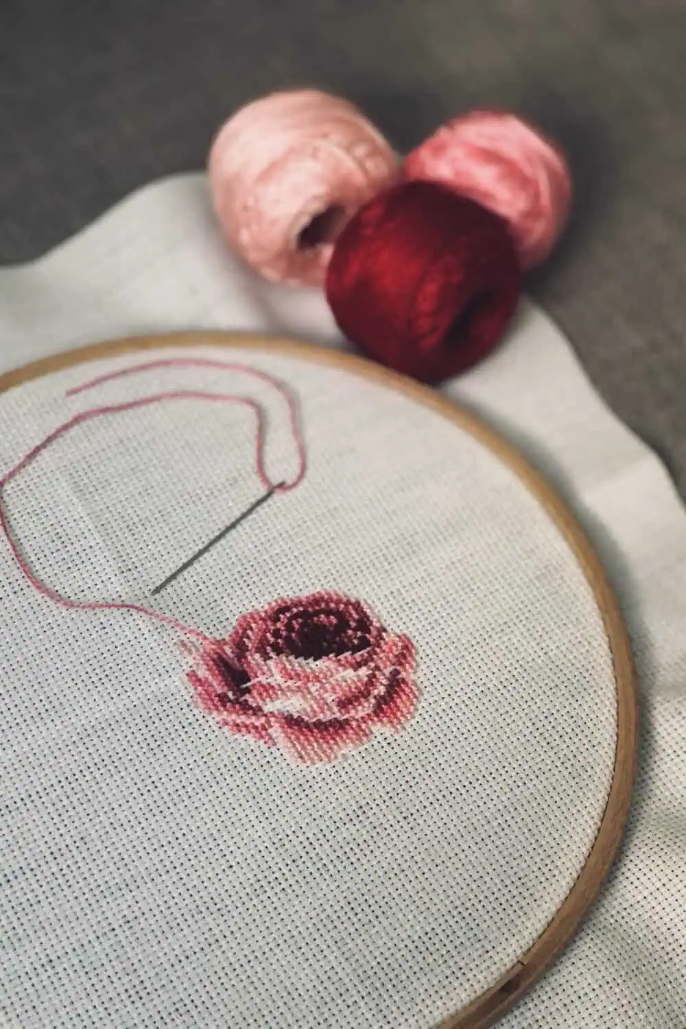 An embroidery hoop with a rose design mid-stitch, a threaded needle resting on the fabric beside two skeins of thread.