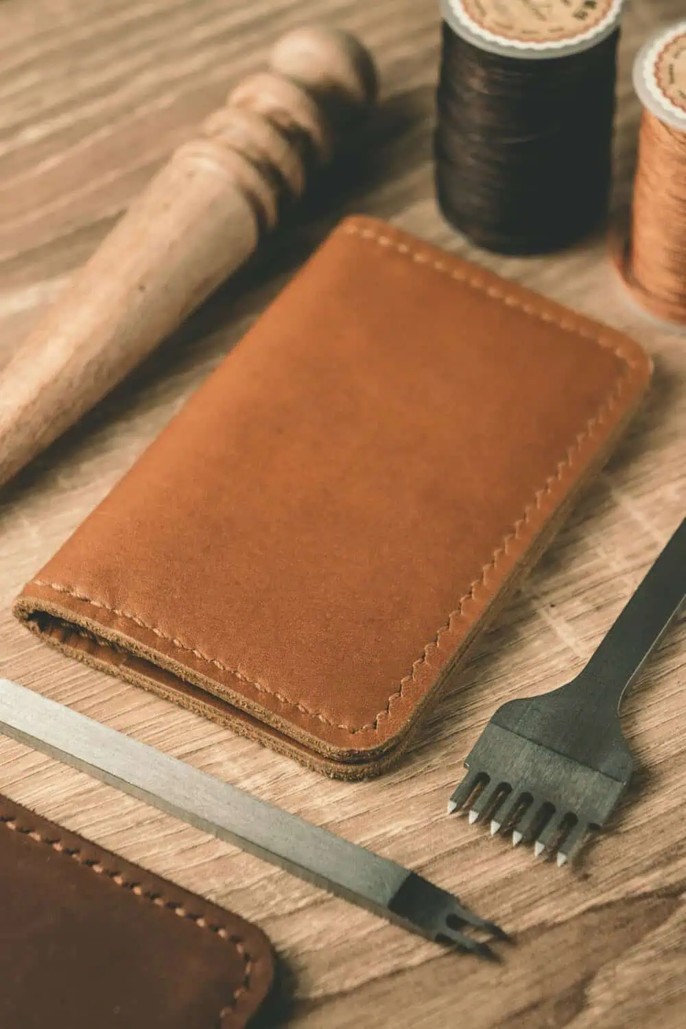 A hand-stitched tan leather wallet resting on a workbench next to leather crafting tools and thread spools.