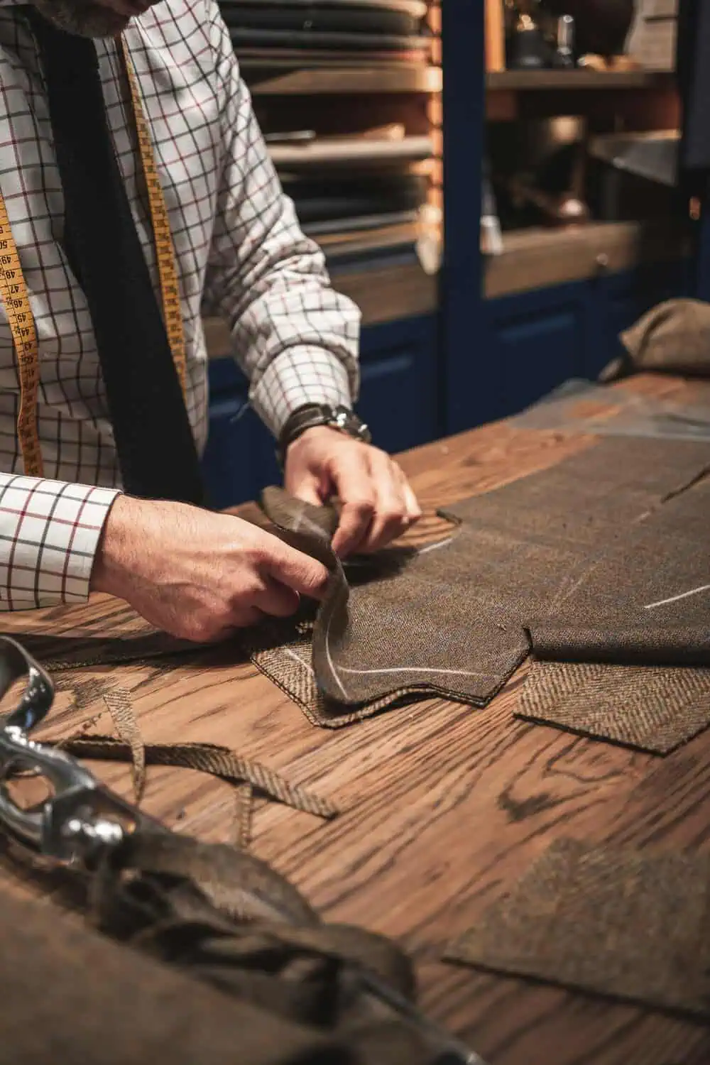 A tailor's hands pinning tweed fabric on a wooden workbench, with a measuring tape draped over his shoulder.