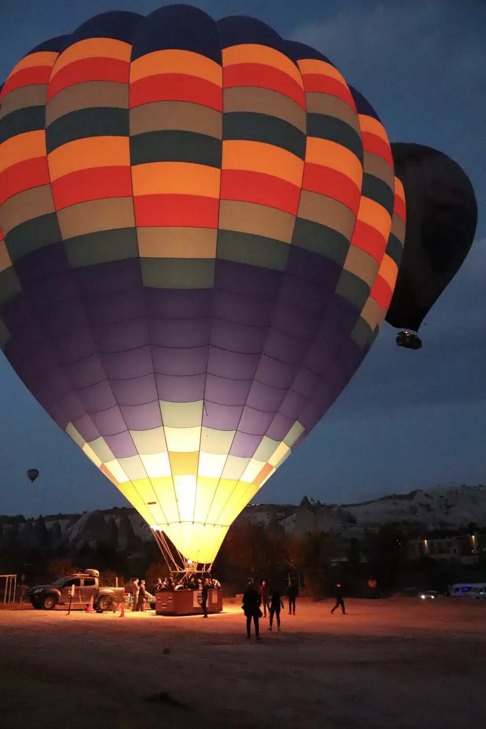 A large striped hot air balloon glowing from its burner flame at dusk, with a second balloon rising behind it.