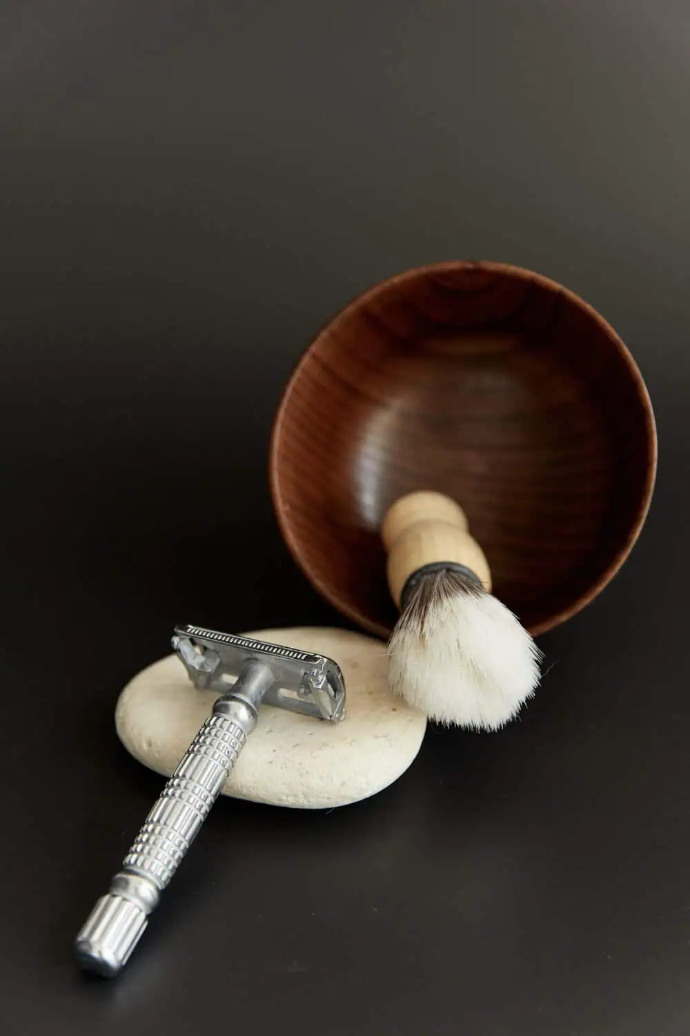 A safety razor and a shaving brush with a wooden handle resting against a wooden bowl on a dark background.