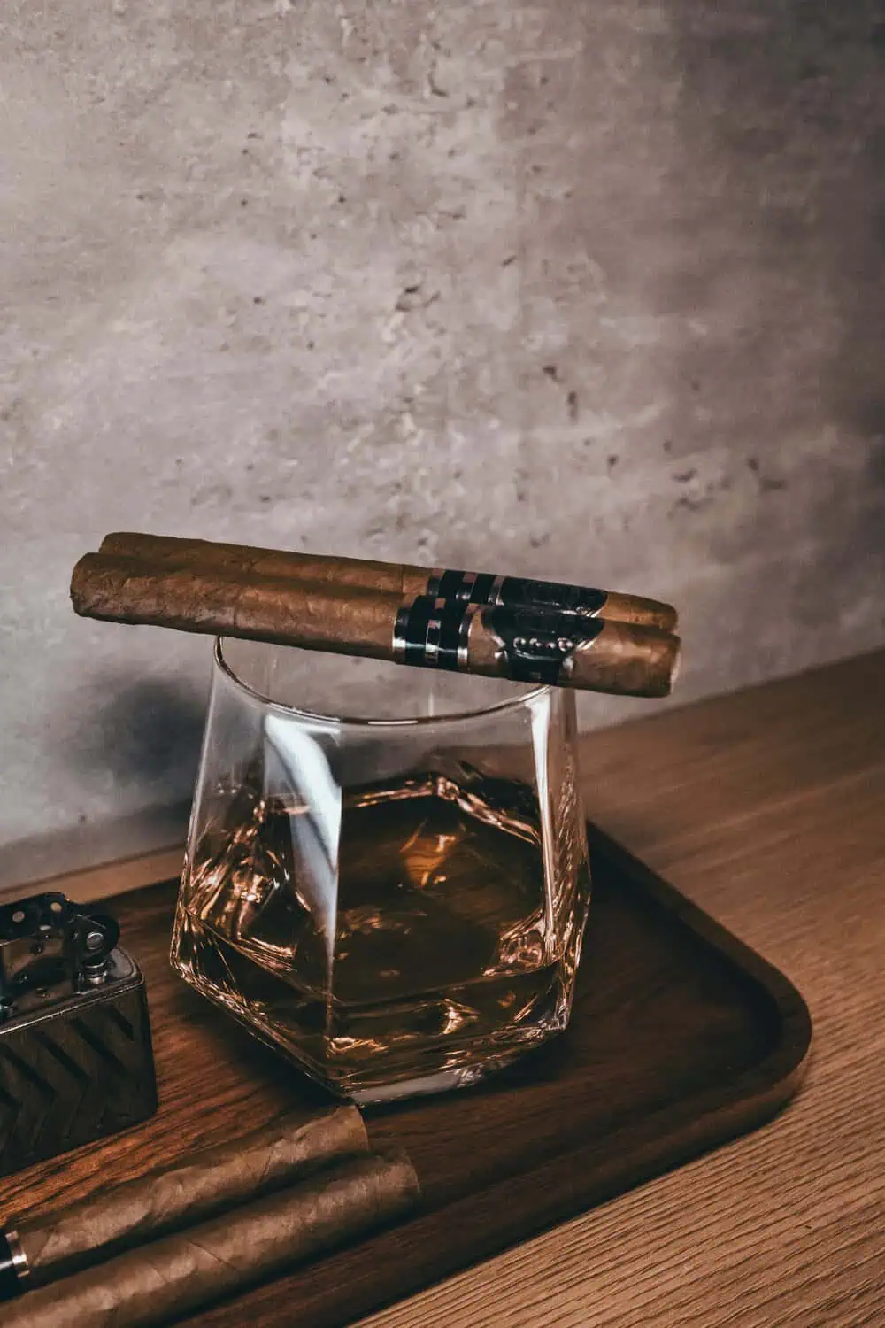A cigar resting across a cut-crystal whiskey glass on a wooden tray, with a lighter and a second cigar nearby.