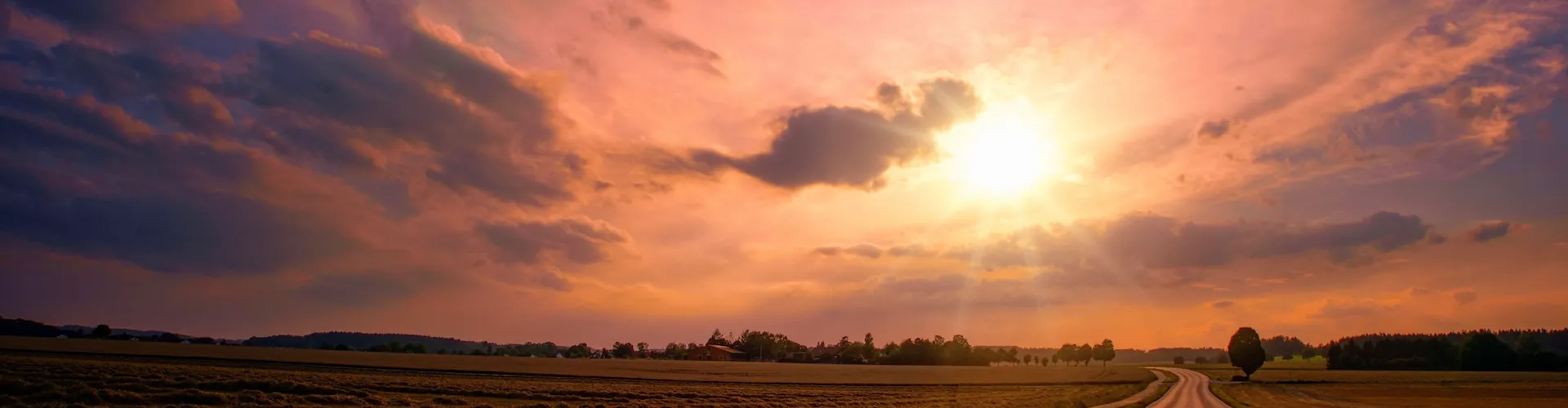 A wide view of a country road at sunset, with sunlight breaking through clouds over open fields.