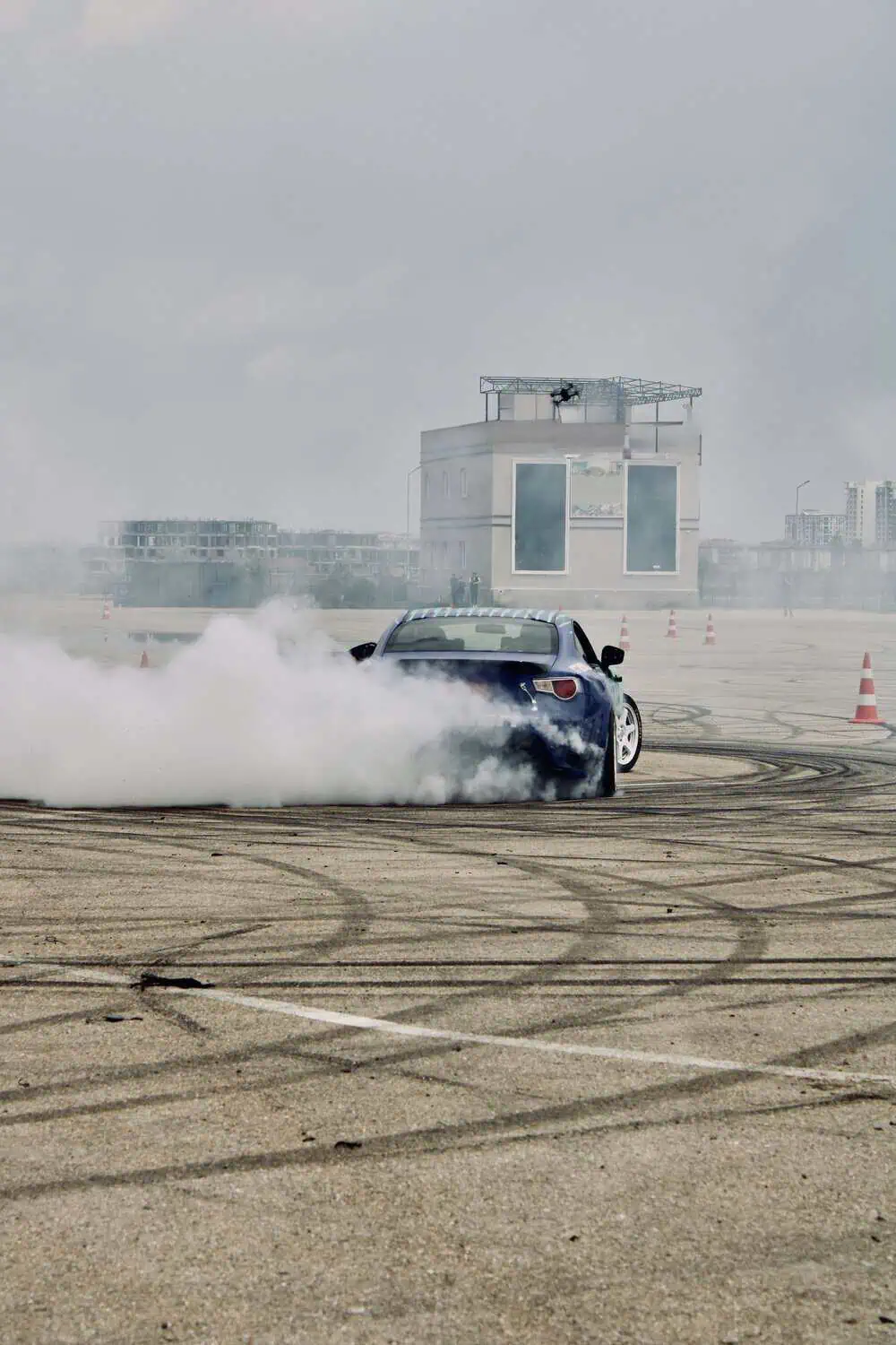 A car drifting sideways on a paved lot, throwing up a large cloud of tire smoke near orange traffic cones.