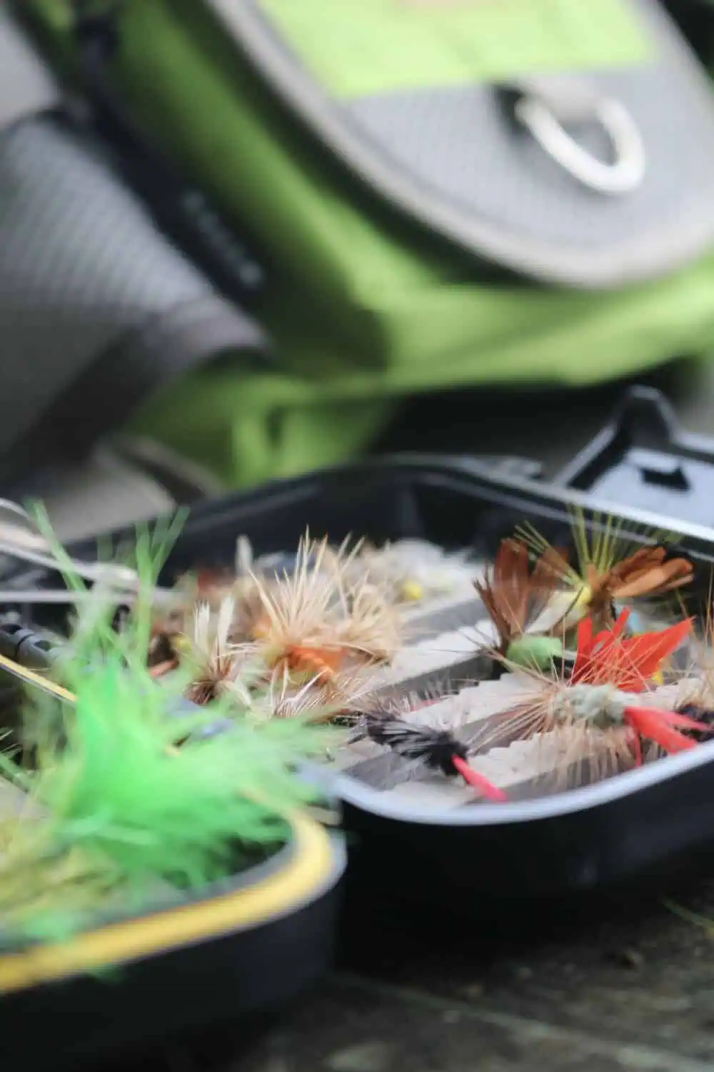 An open tackle box filled with colorful fly fishing lures next to a rod and fishing pack.