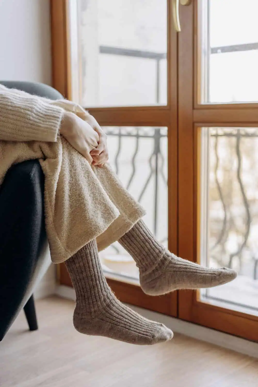 A person's legs in knit compression socks tucked under a cream blanket while resting near a sunlit window.