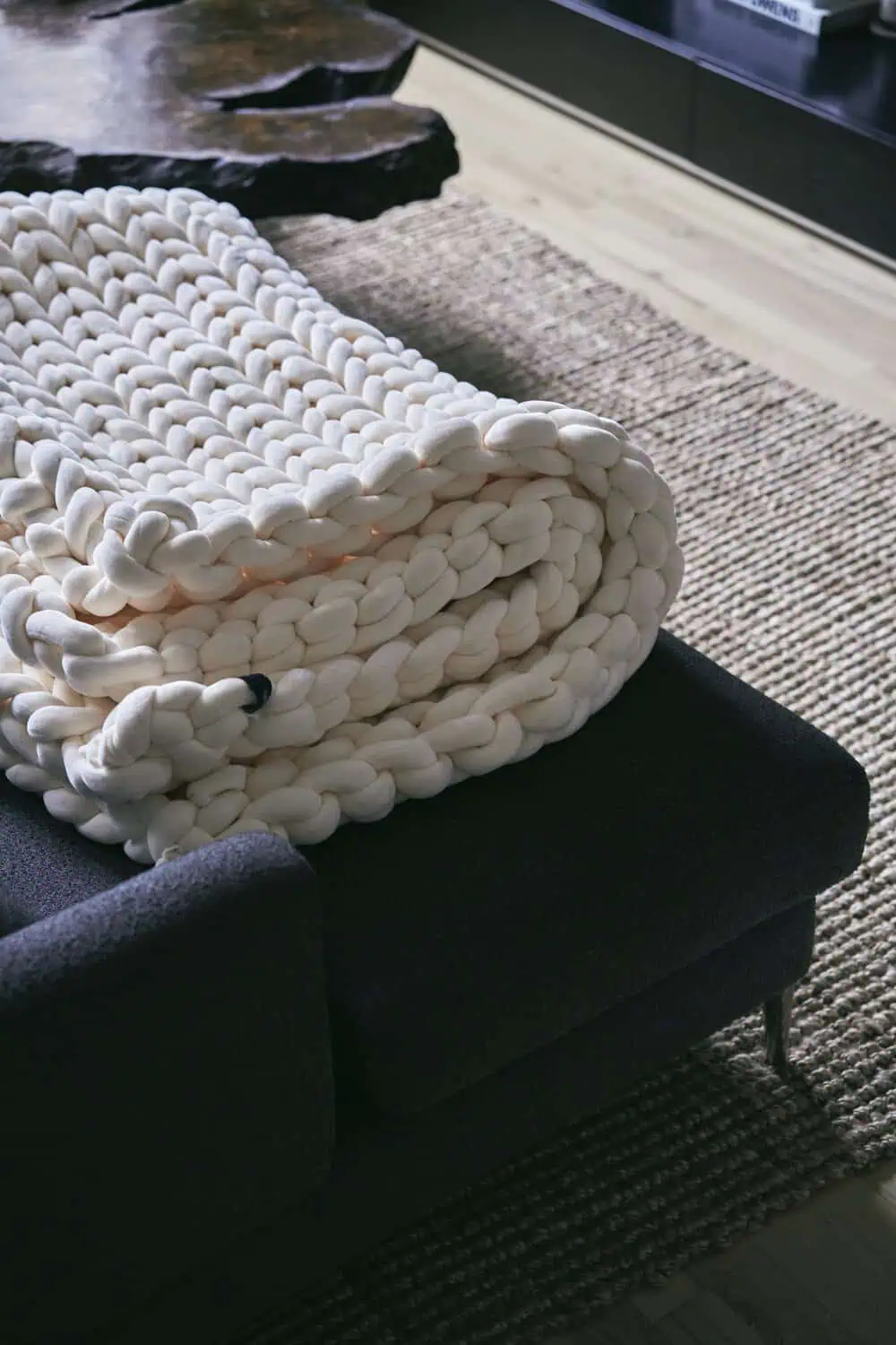 A thick, chunky knit blanket rolled up on a dark ottoman beside a low wood coffee table.