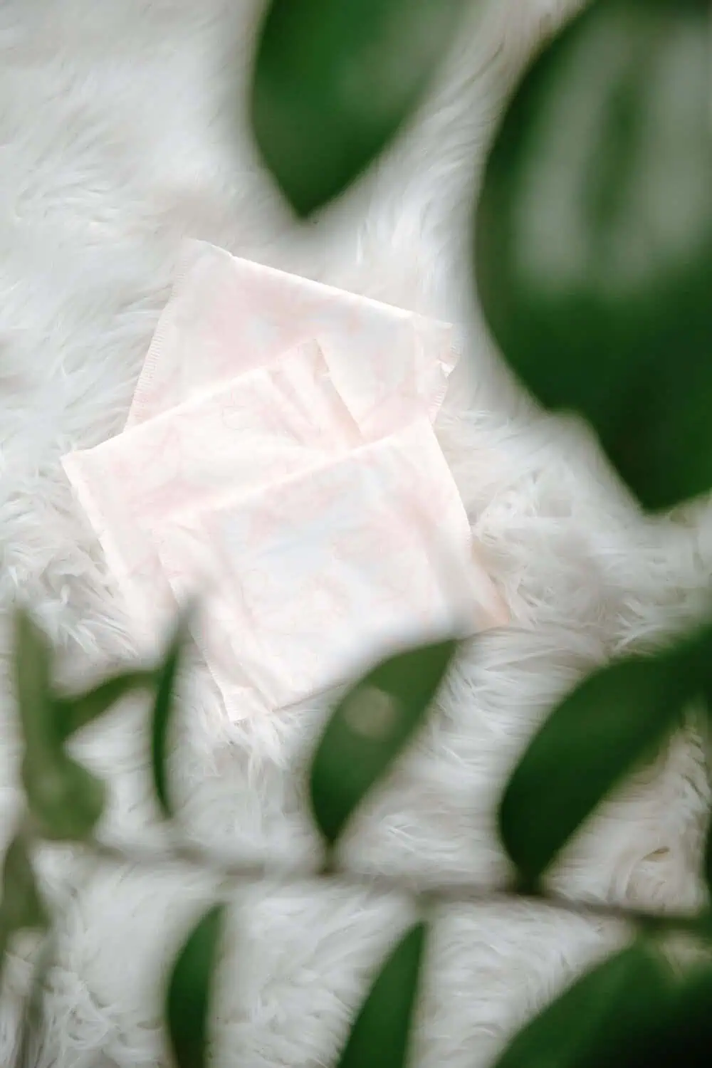 Two folded pale pink postpartum ice pack pads resting on a white faux fur blanket with green leaves in the foreground.