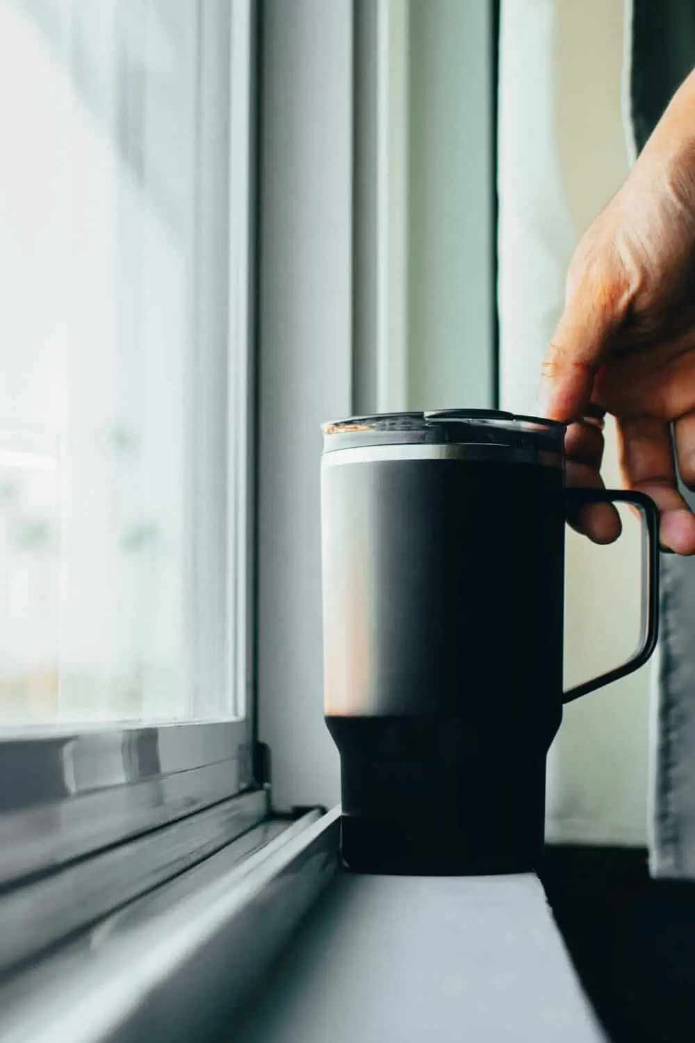 A hand lifting a black insulated tumbler with a handle from a window sill.