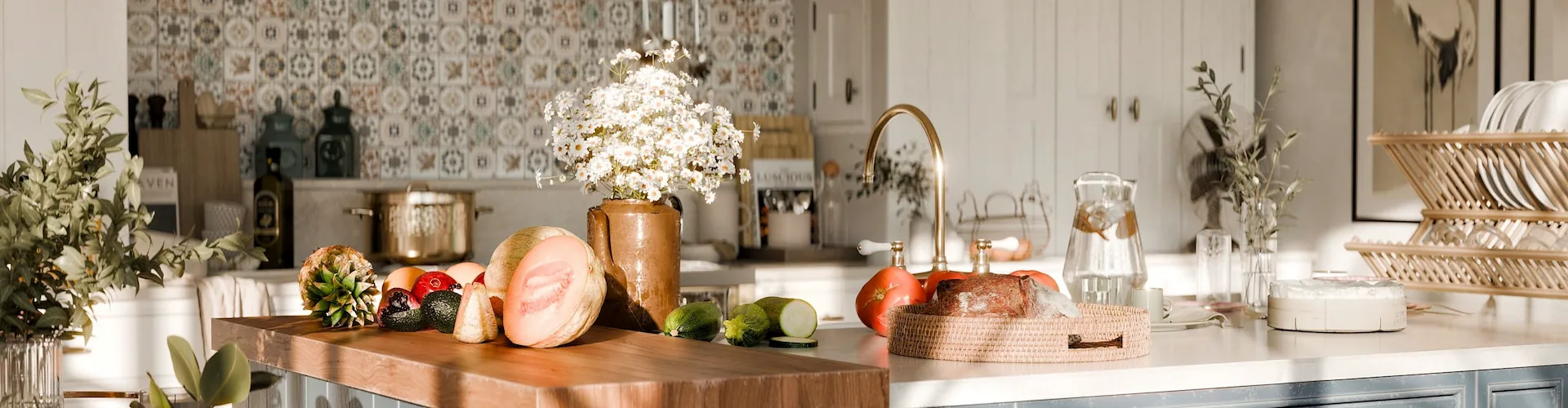 A bright kitchen counter with fresh produce, flowers in a vase, and a gold faucet in warm morning light.