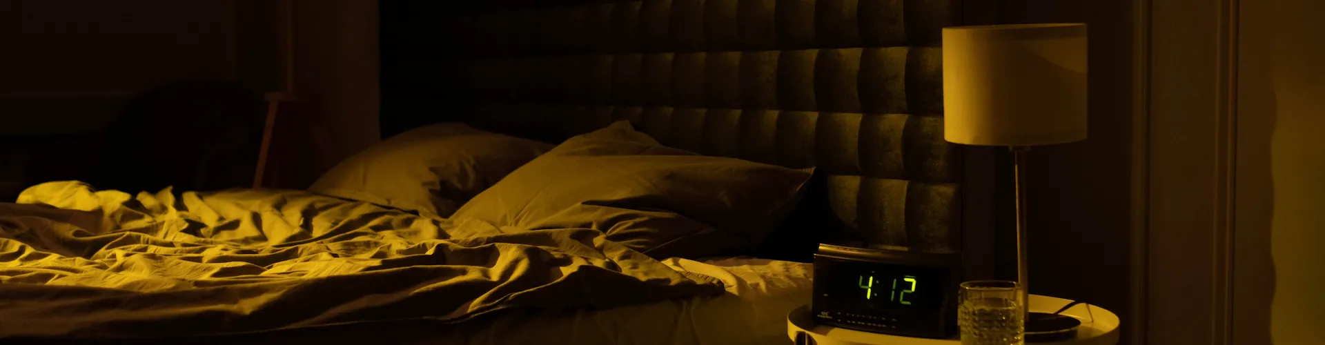 A dimly lit bedroom at night with an unmade bed, a glowing digital clock reading 4:12, and a warm bedside lamp.