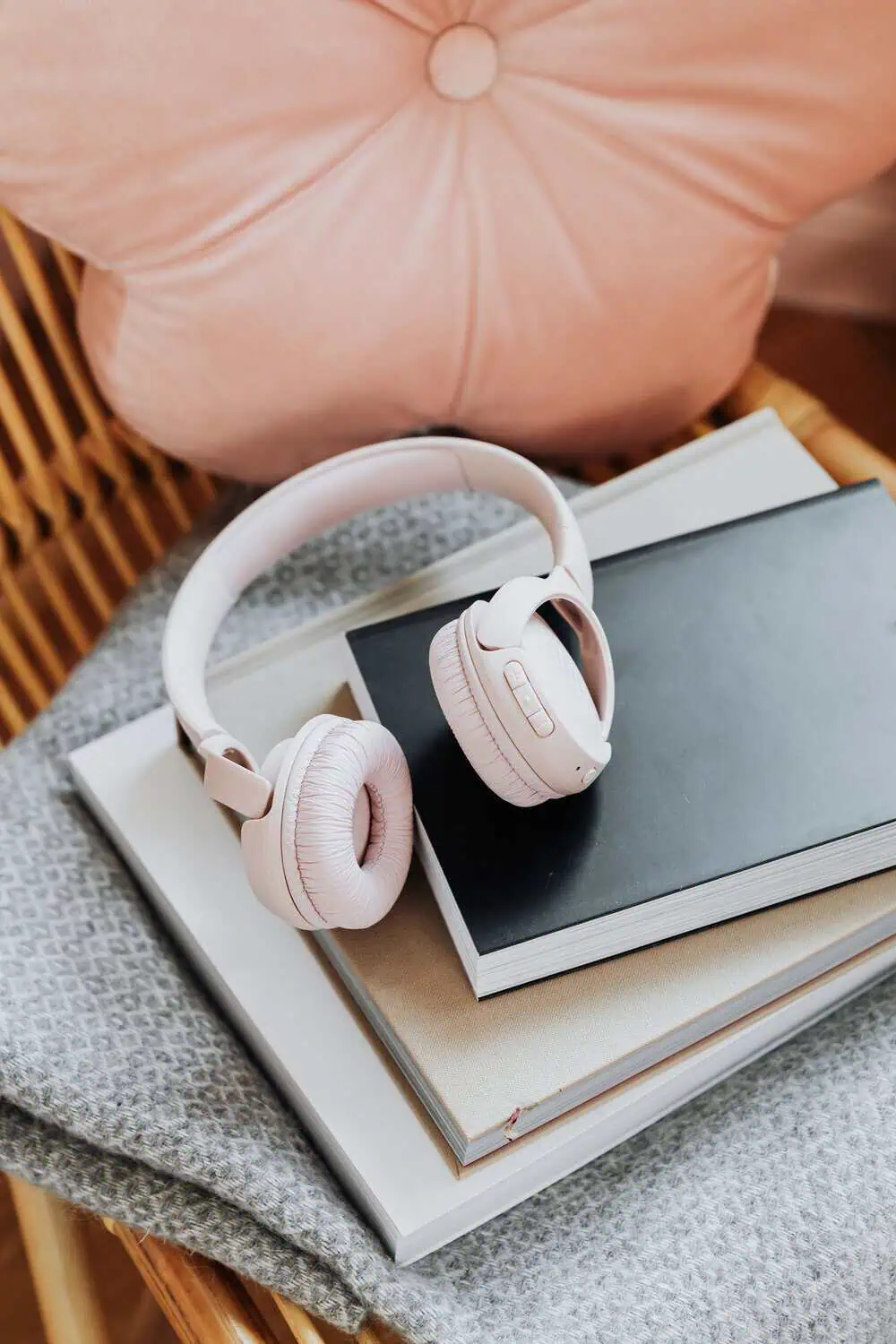 Blush pink over-ear headphones resting on a stack of books next to a pink cushion.
