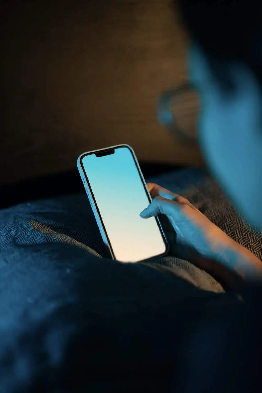 A hand touching the glowing screen of a phone while sitting on a couch in a dark room at night.
