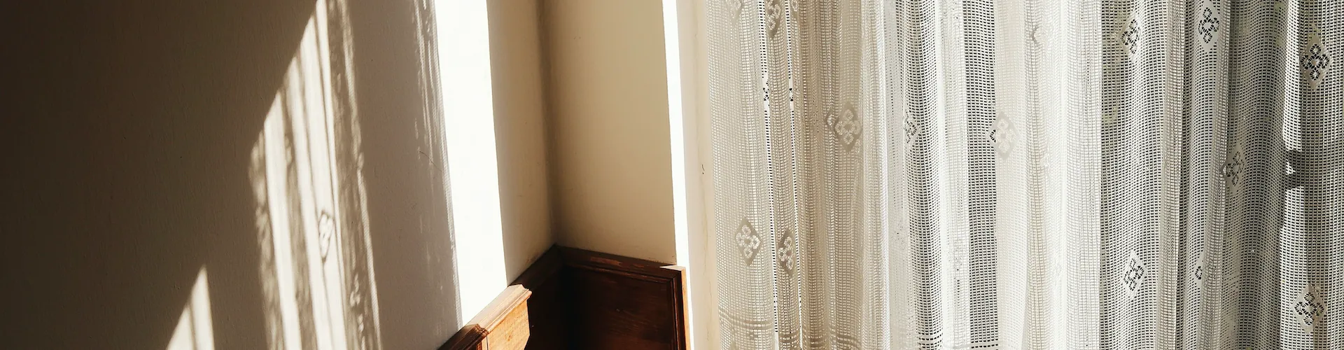 Afternoon sunlight streaming through a sheer lace curtain into a quiet room corner.