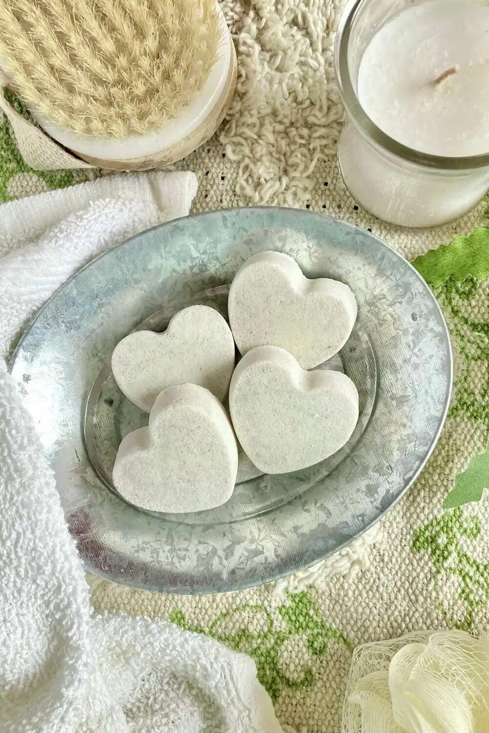Four heart-shaped shower steamer tablets arranged on a metal tray beside a lit candle and a body brush.