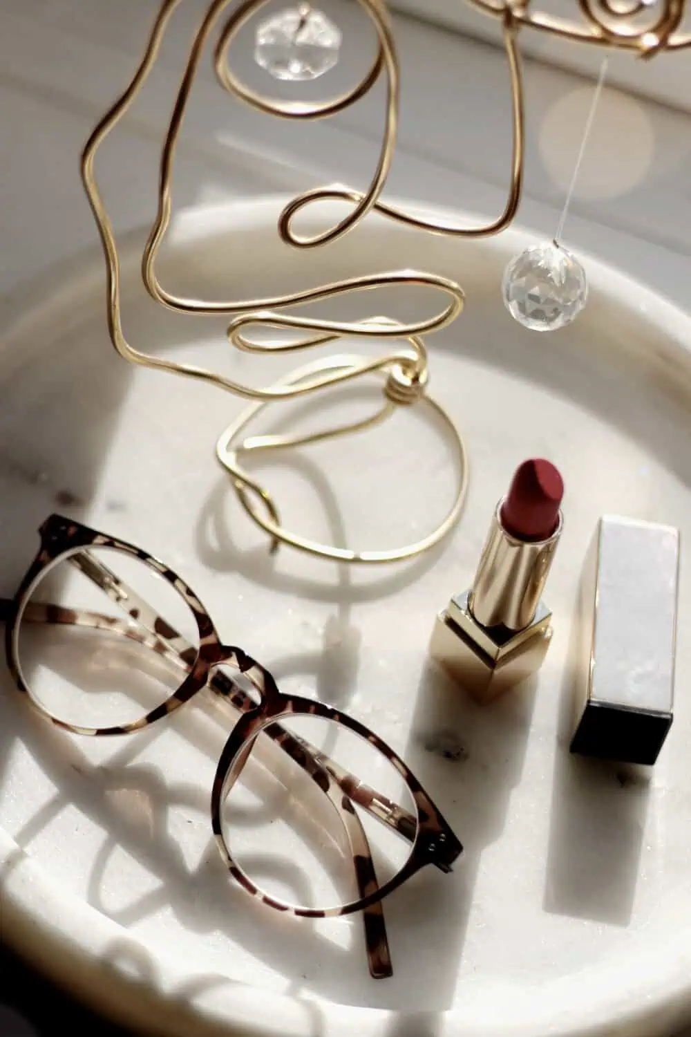A round marble tray holding gold wire jewelry, tortoiseshell glasses, and a red lipstick in sunlight.