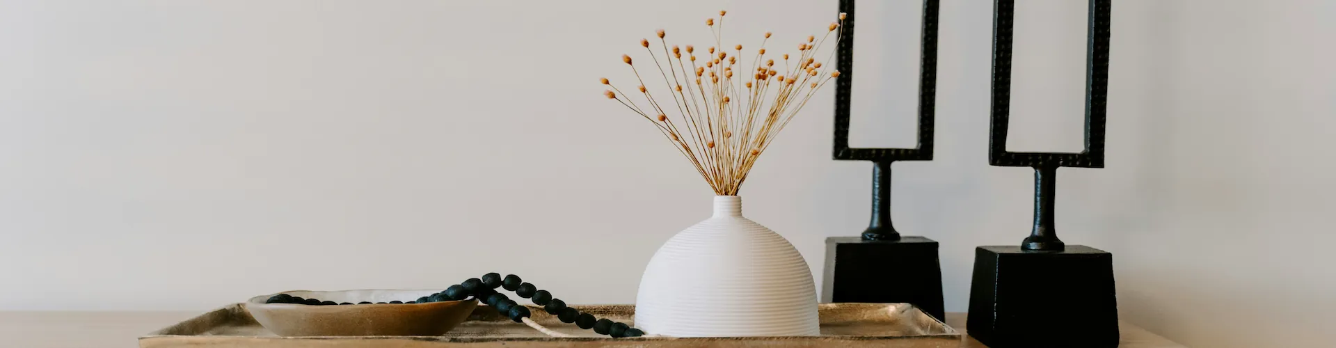 A wide styled tabletop with a white bud vase of dried flowers, a bowl of dark beads, and two dark frame stands.