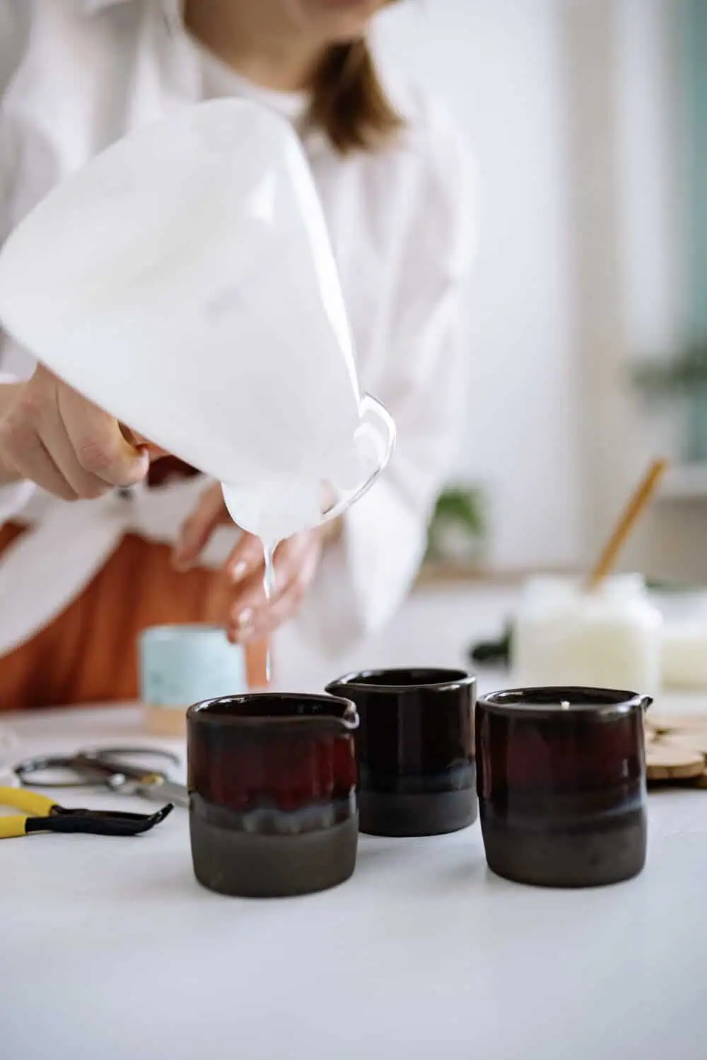 A person pouring melted wax from a glass pitcher into three dark ceramic candle vessels on a table.