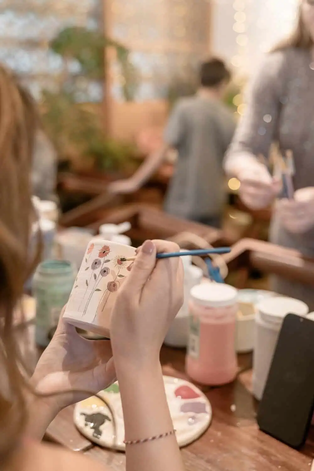A hand painting small flowers onto an unglazed ceramic mug at a pottery painting studio table.