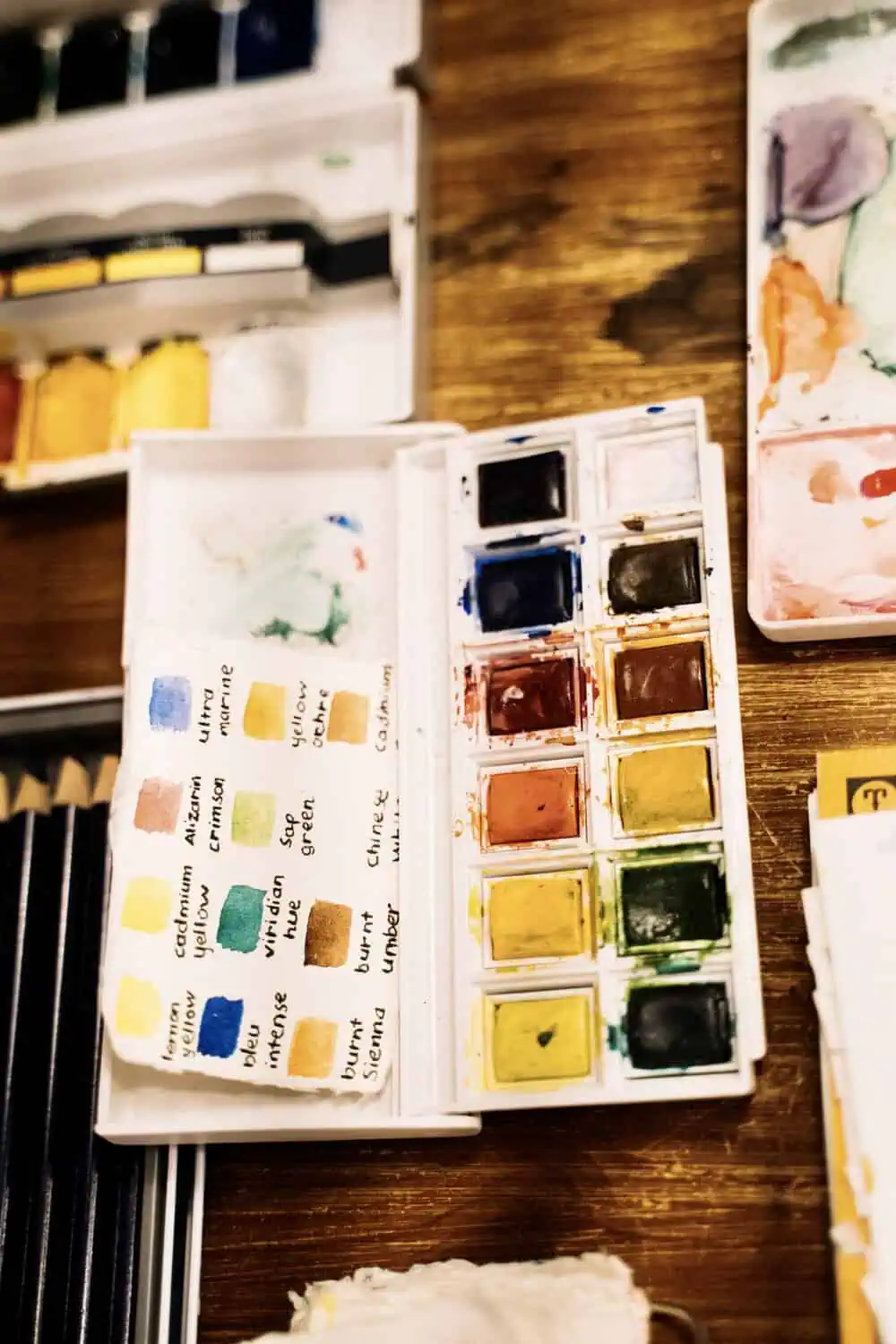 An open watercolor palette with hand-labeled pigment swatches next to a case of colored pencils.