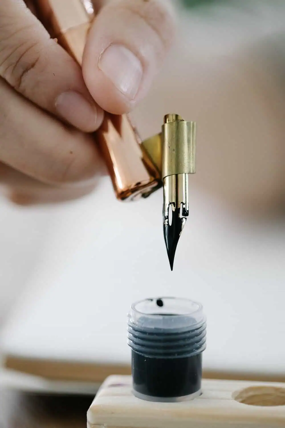 A hand dipping a gold fountain pen nib into a small bottle of black ink.