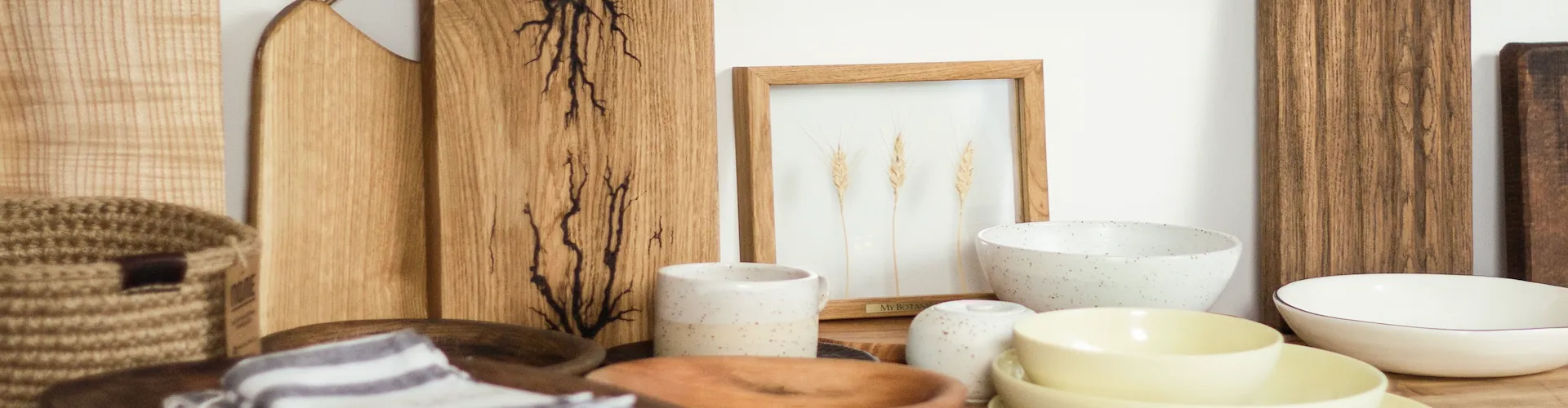 A styled arrangement of wooden cutting boards, speckled ceramic bowls, and a small framed print of dried wheat stalks sits together on a table.