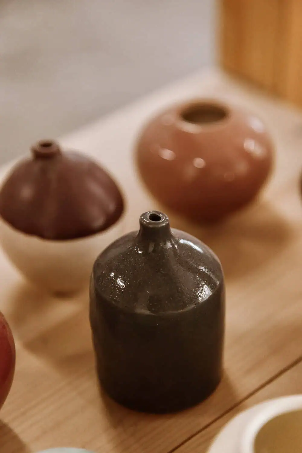 A small dark ceramic bud vase in focus on a wood table, with two more bud vases blurred behind it.