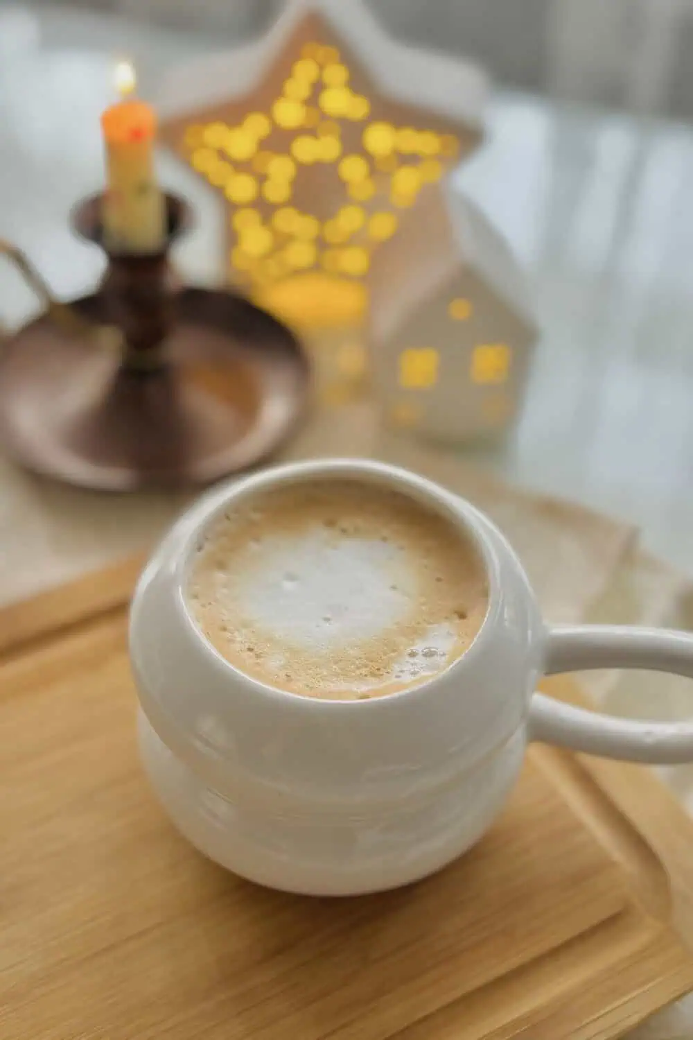 A white ceramic mug of latte with foam on top, a lit candle and string-light houses blurred in the background.