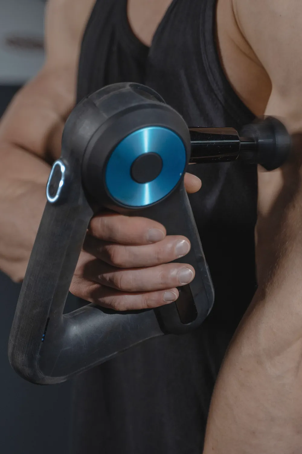 A hand holding a black percussion massage gun with a blue accent against a dark tank top.