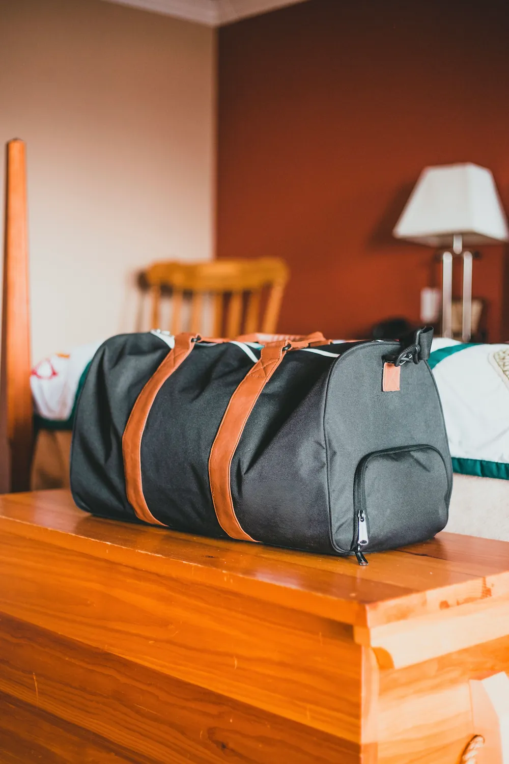A black canvas duffel bag with tan leather straps sitting on a wooden dresser in a bedroom.