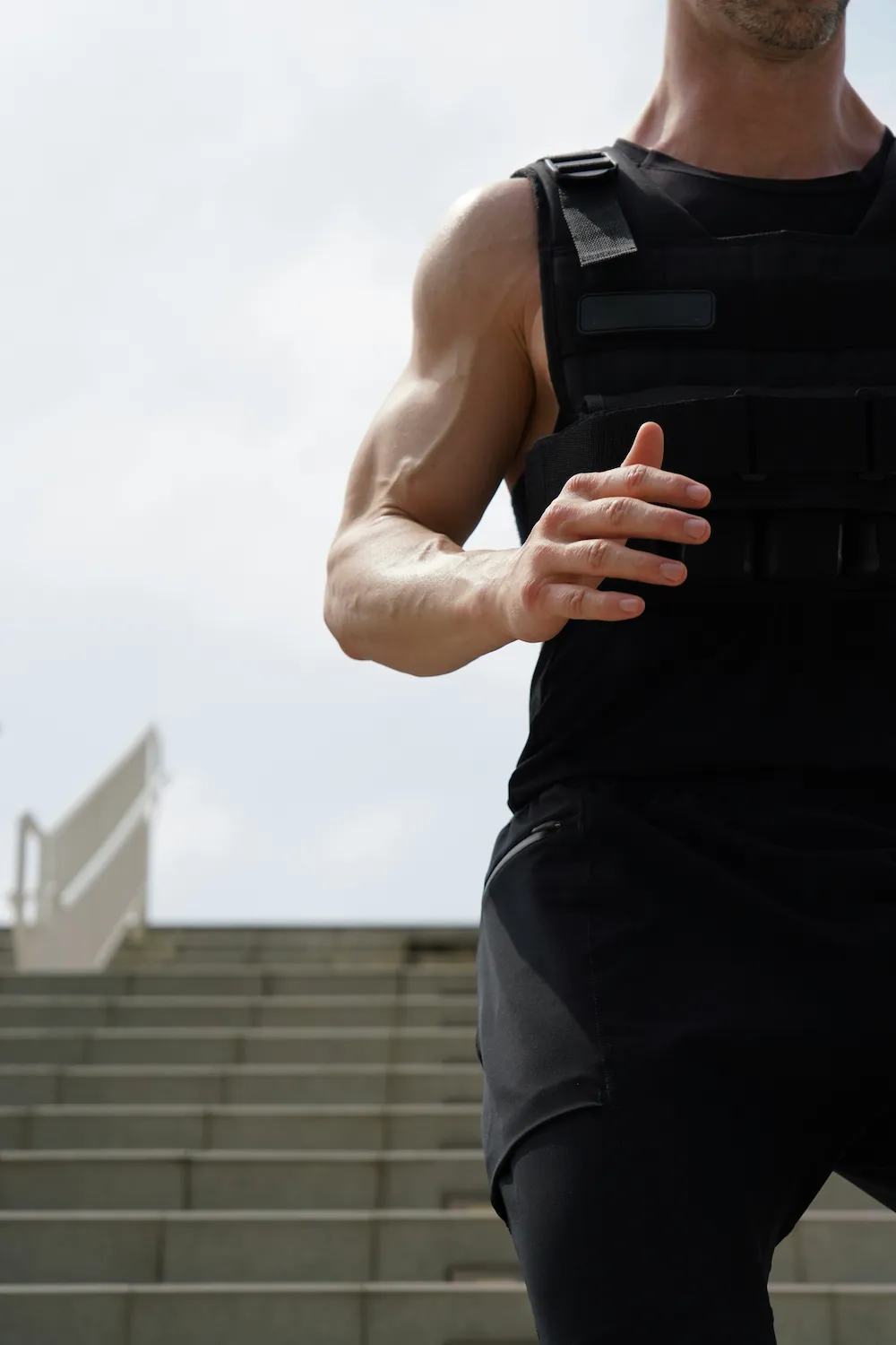 A muscular man wears a black weighted vest while standing outdoors near a staircase.