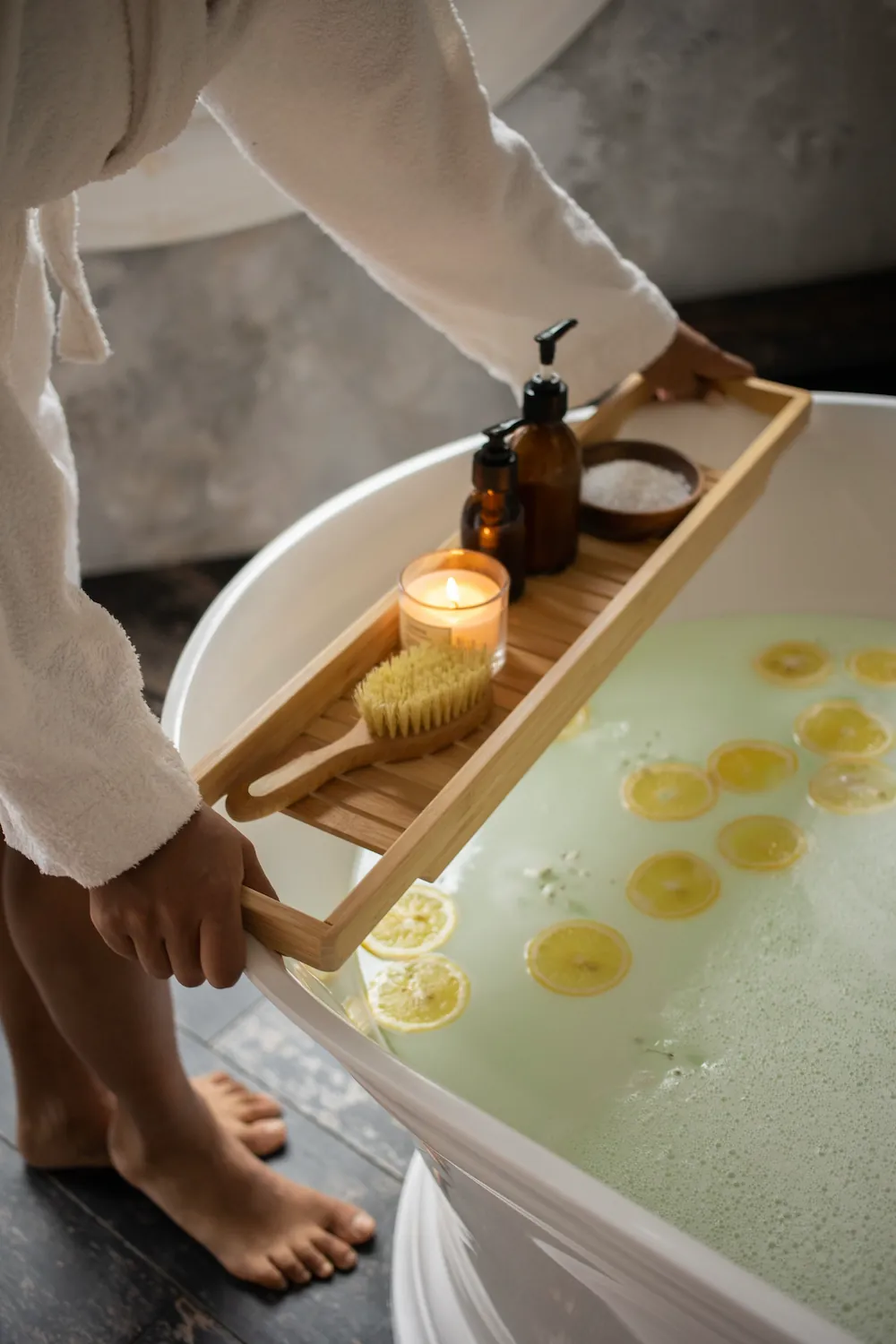 A wooden bath tray holding a candle, bath salts, oils, and a brush balanced across a tub filled with lemon slices.