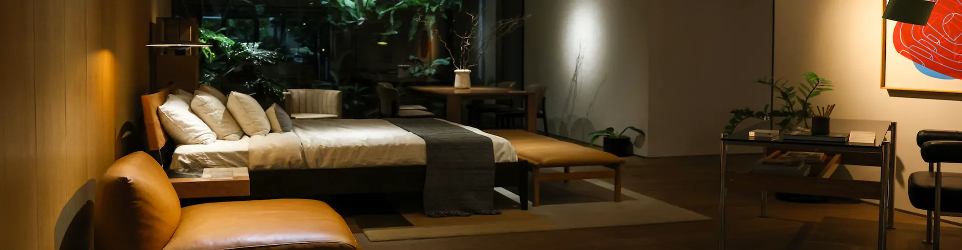 A dimly lit bedroom with a made bed, leather bench, and lounge chair in a warm evening glow.