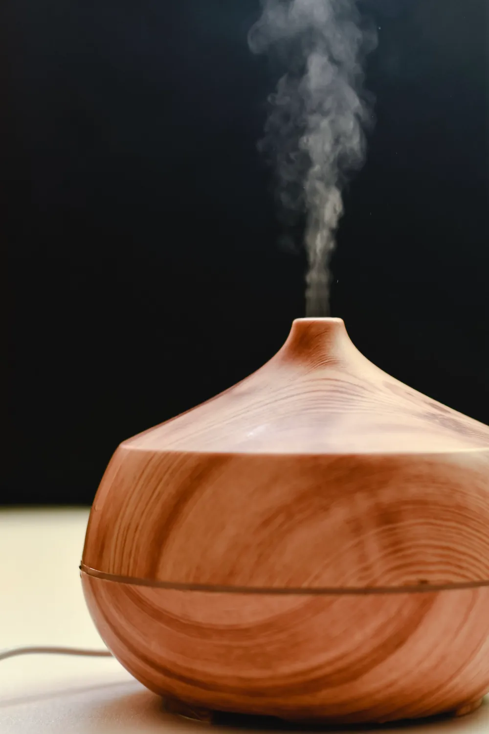 A close-up of a wood-grain essential oil diffuser releasing a stream of visible steam against a dark background.