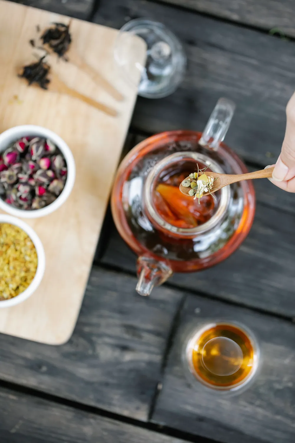 A glass teapot steeping tea next to bowls of dried rose buds and herbs, with a spoon adding chamomile.