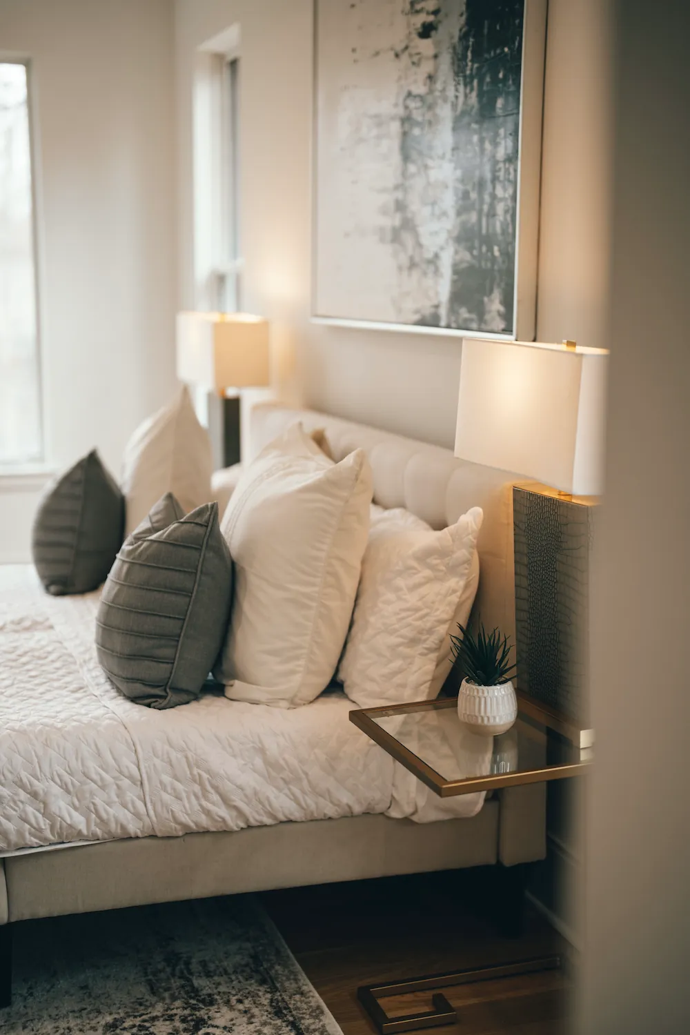 A made bed with layered white and grey pillows against an upholstered headboard, lit by a bedside lamp.