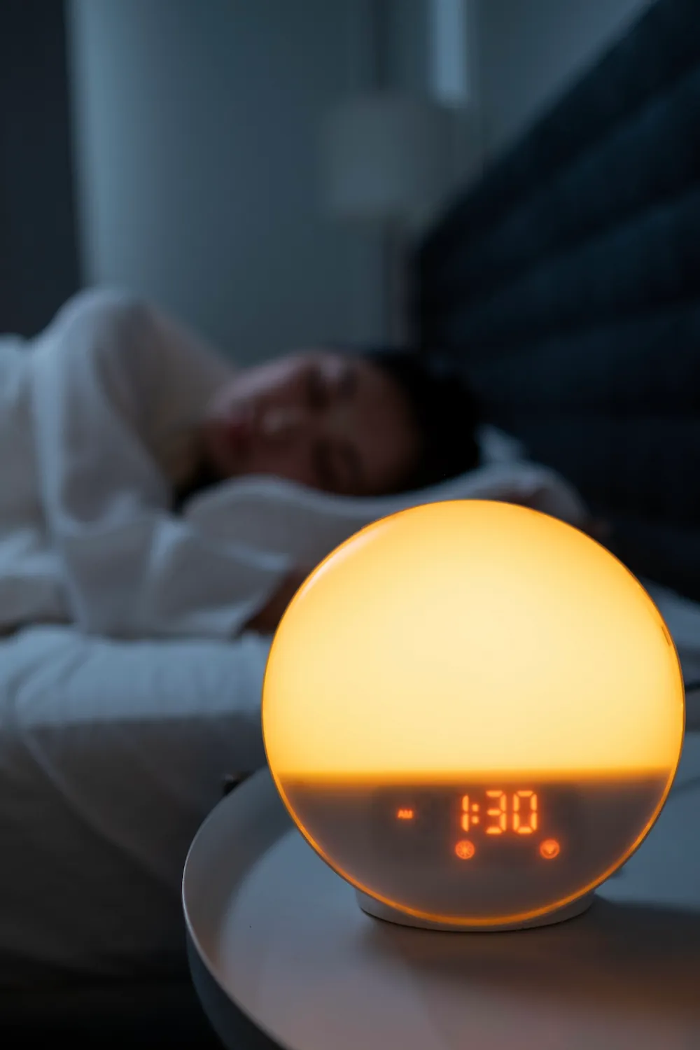 A round sunrise alarm clock glowing orange on a nightstand while someone sleeps in the background.