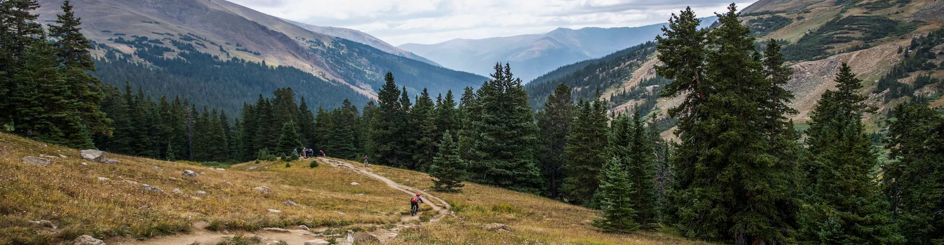 A mountain trail winds through pine trees with distant hikers, framed by layered mountain ridges under a partly cloudy sky.