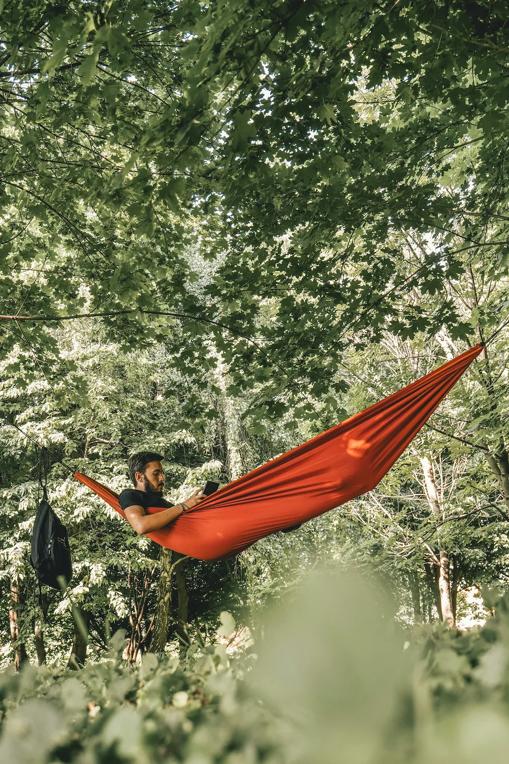 A man lies in an orange camping hammock strung between trees, looking at his phone, with a black backpack hanging nearby.