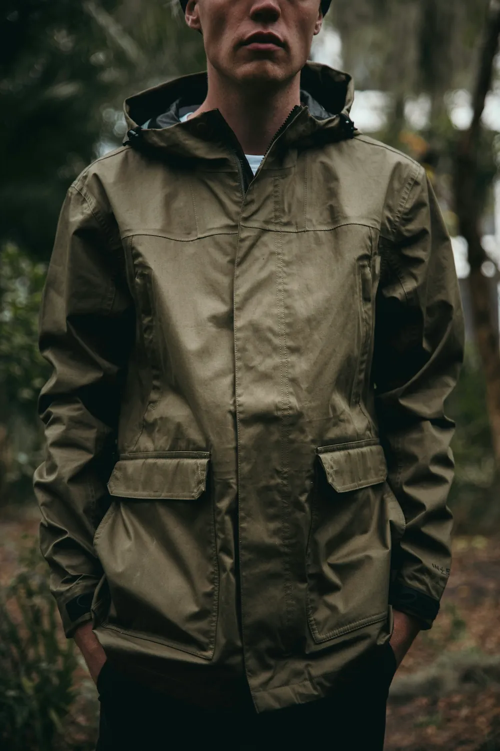 A man wearing an open-collared olive green hooded rain jacket stands among trees, hands in the jacket's pockets.
