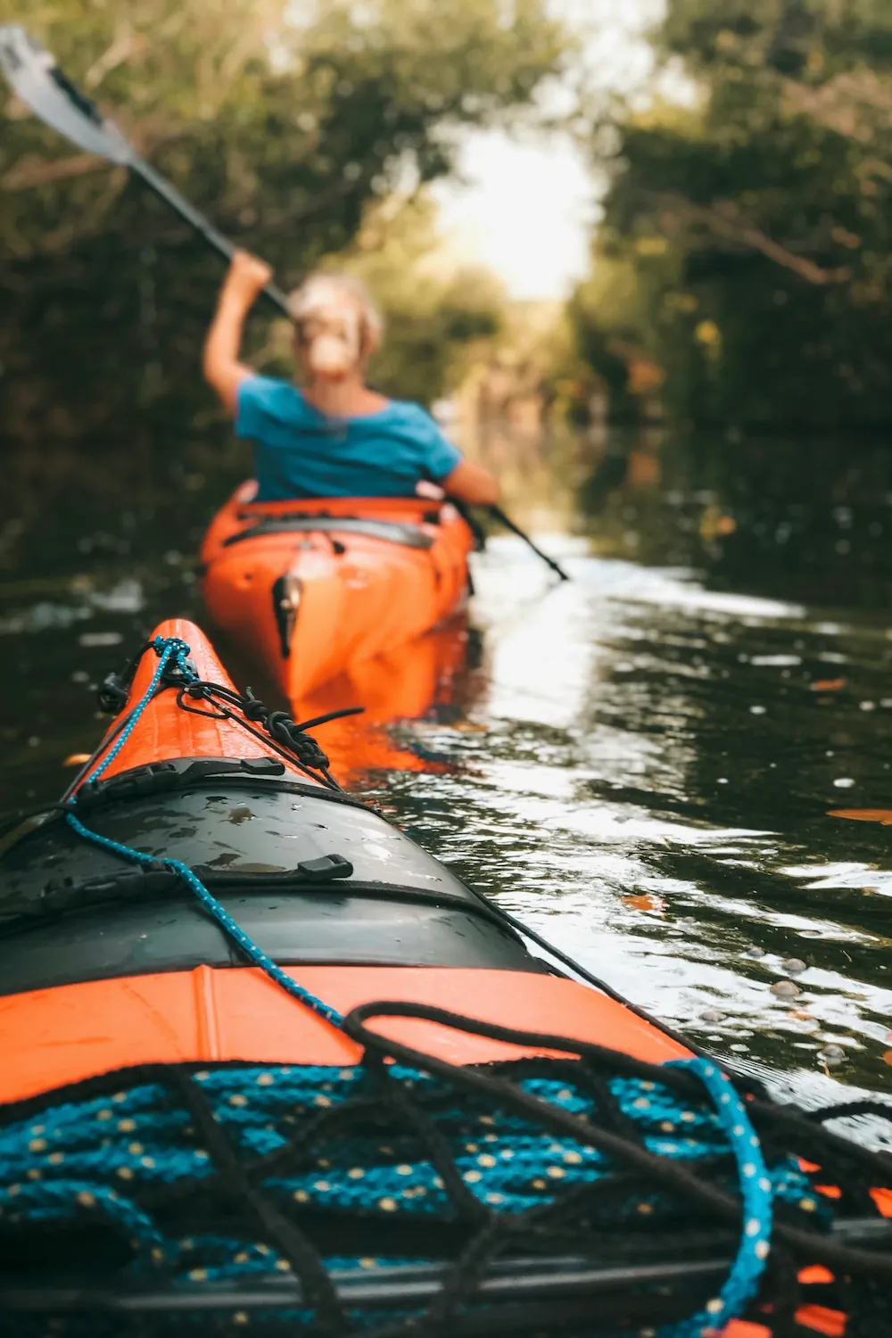 Two kayakers paddling single file down a calm, tree-lined river.