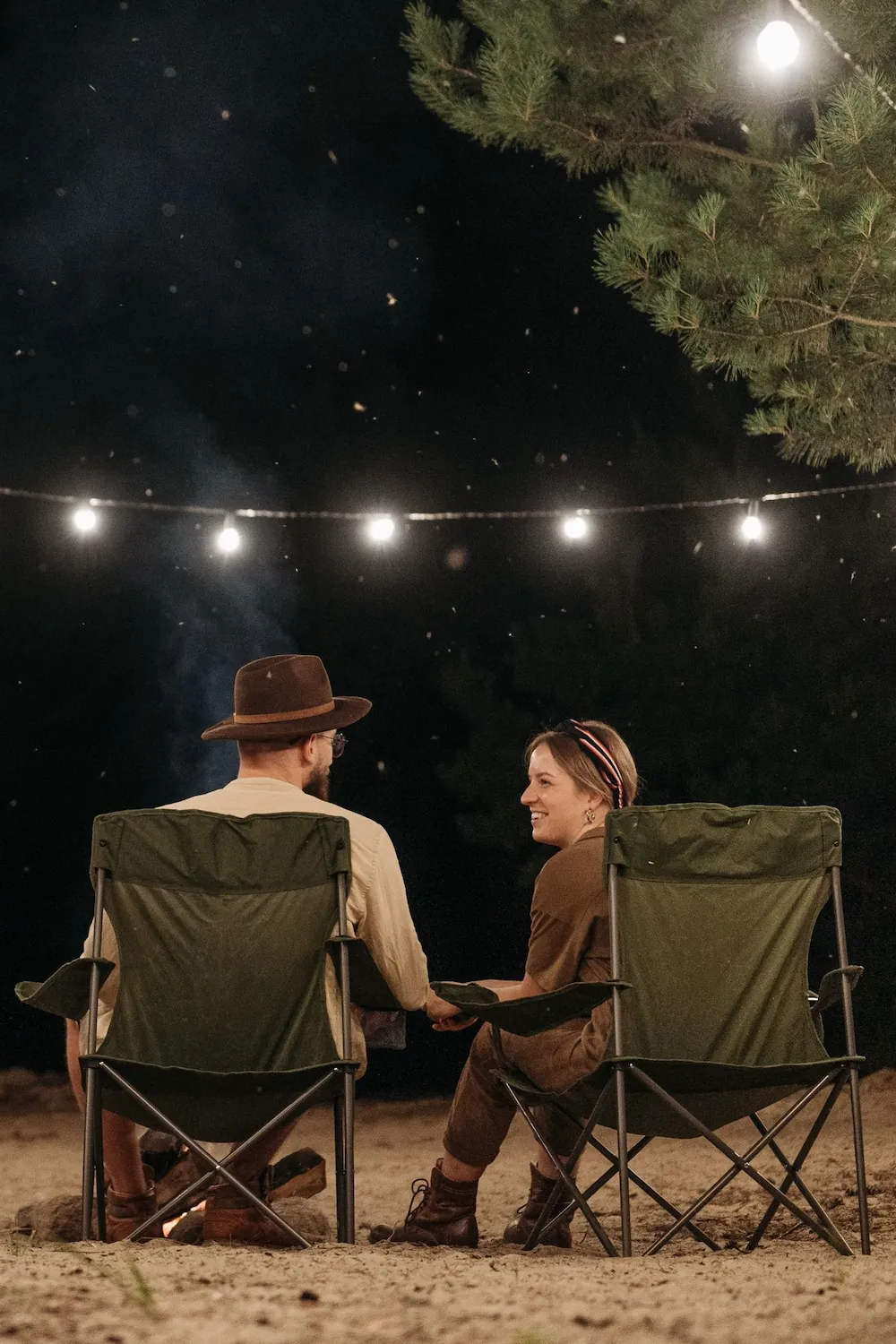 A couple sitting side by side in green camp chairs at night under string lights, holding hands.