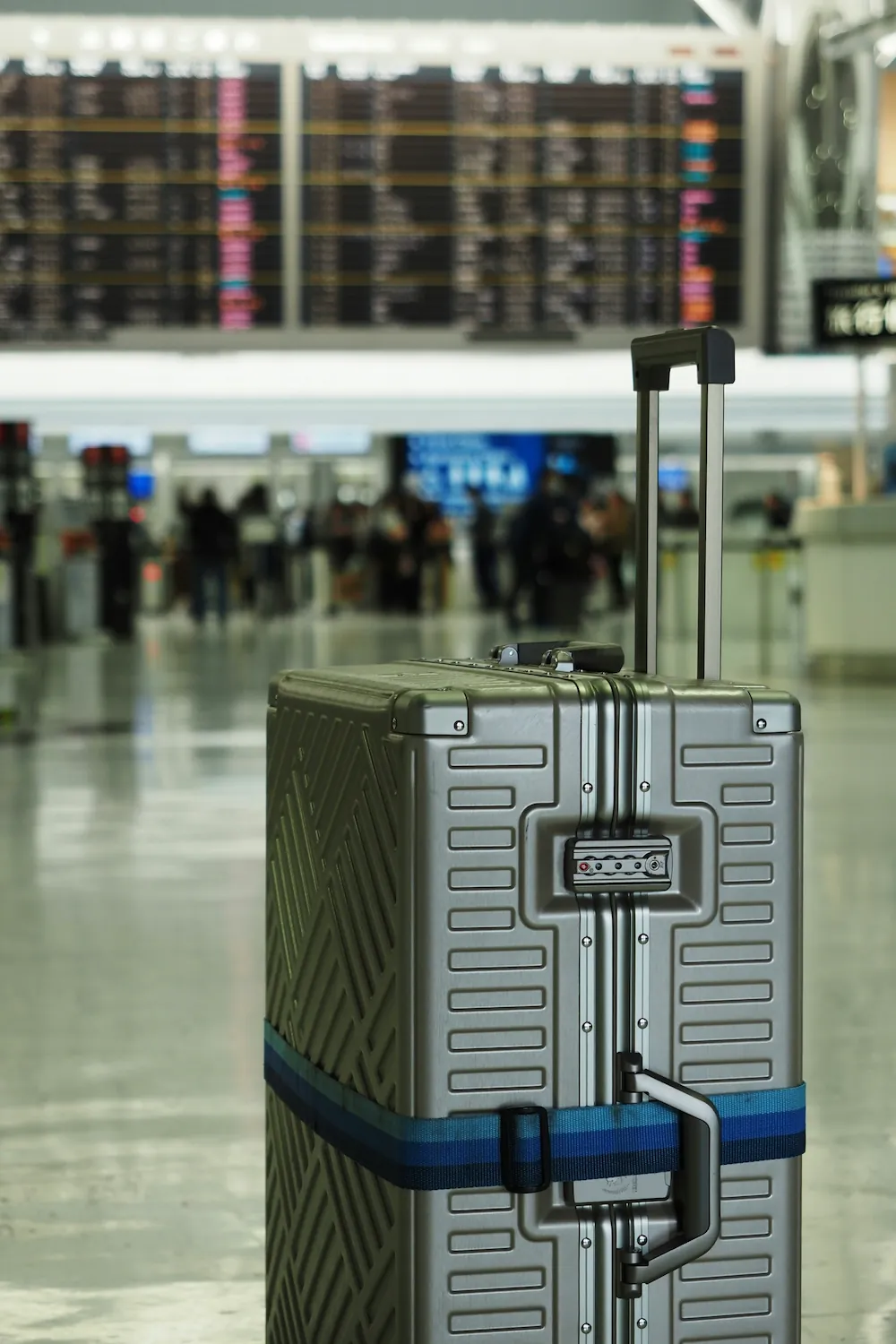 A silver hardside suitcase stands upright in an airport terminal, with a departure board and travelers blurred in the background.