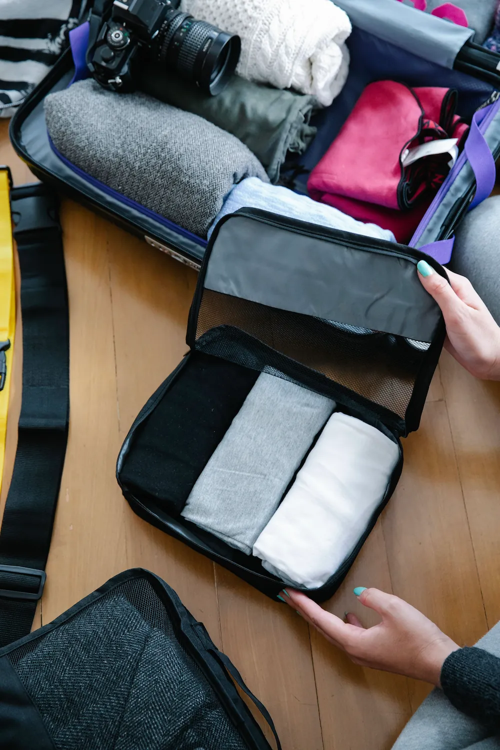 An open suitcase packed with folded clothes and a camera sits next to an open packing cube of neatly rolled shirts, with two hands adjusting the contents.